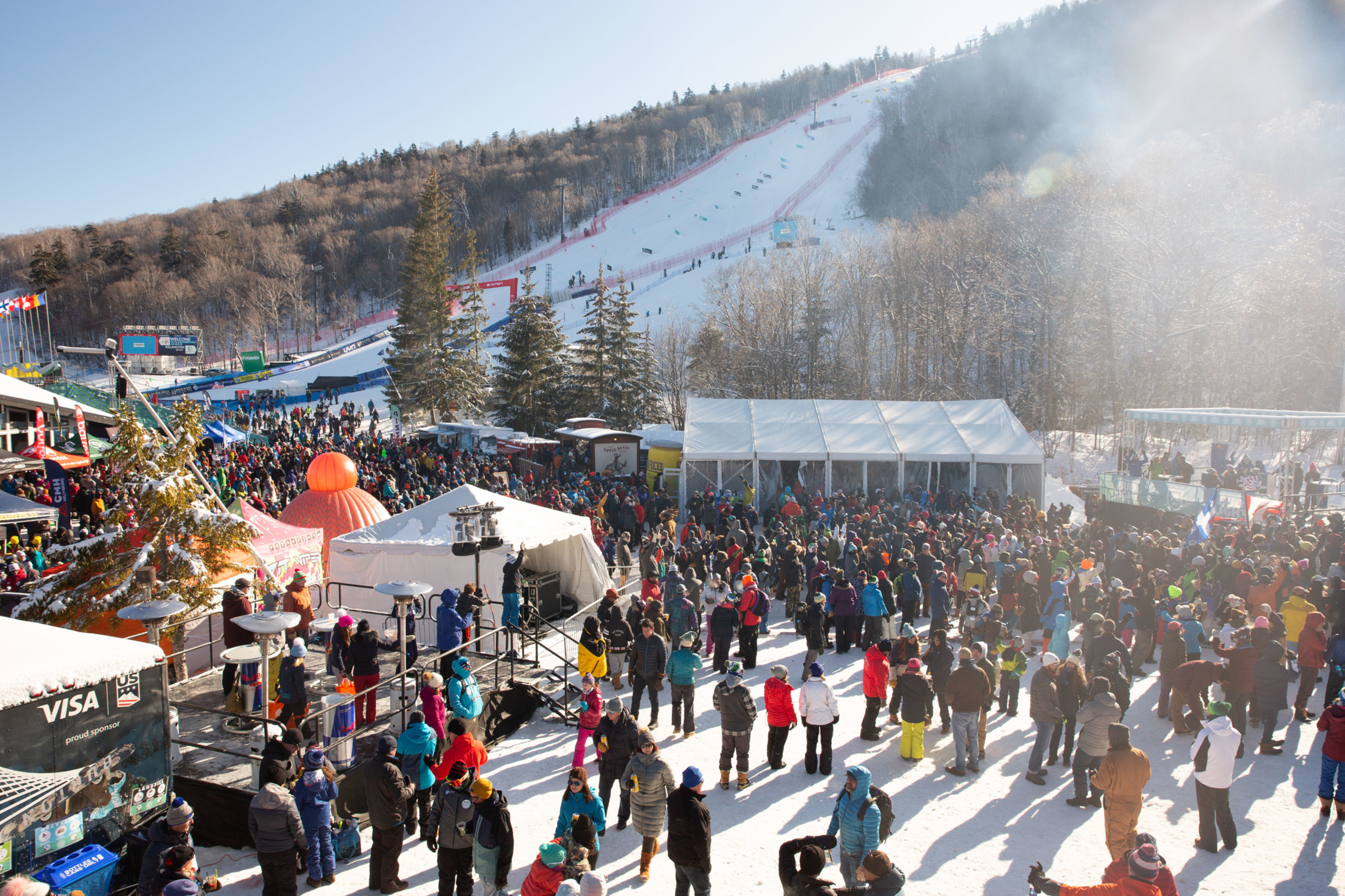 Crowd gathered at a ski resort with snowy slopes and festive tents under a clear sky.