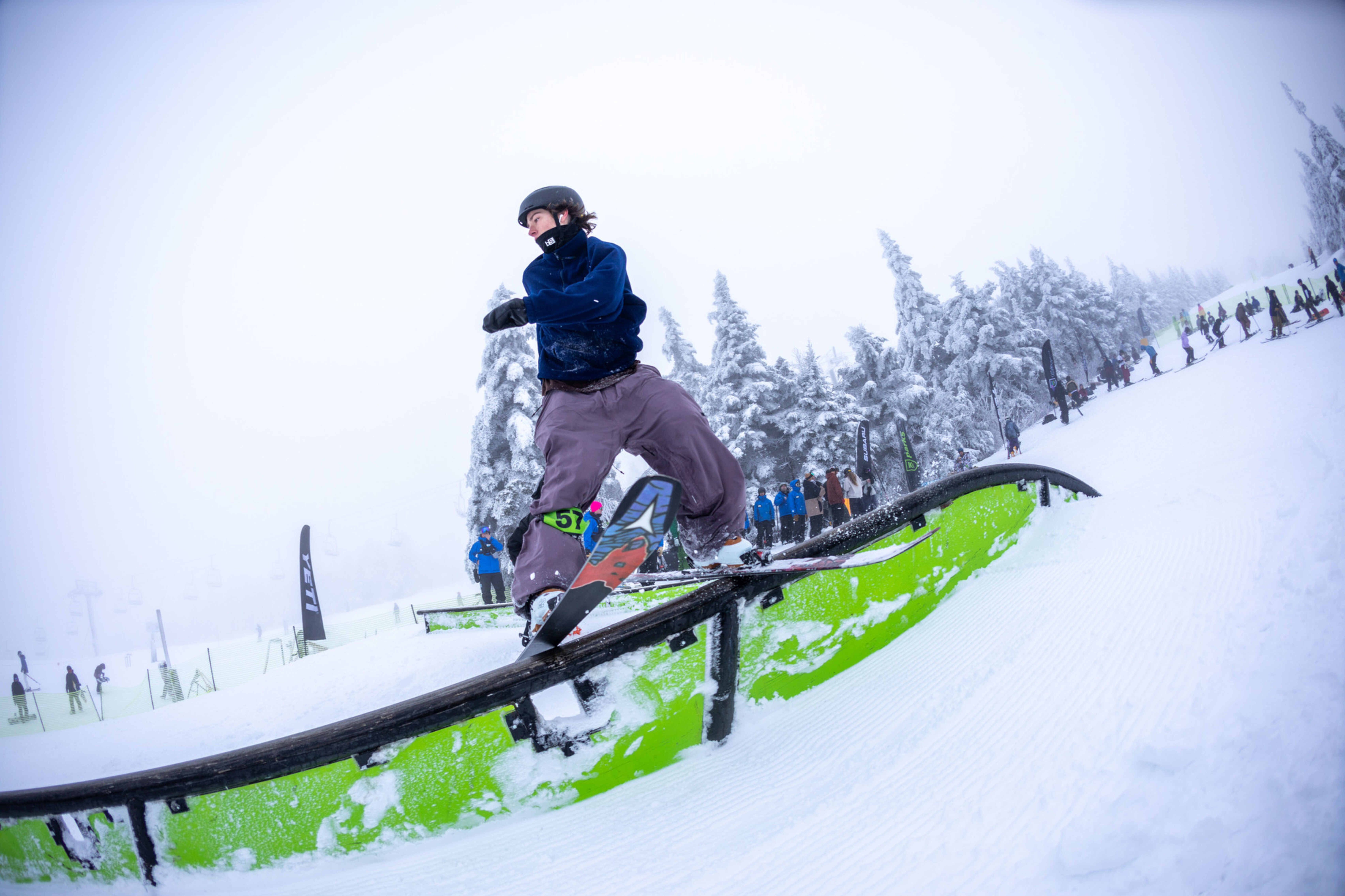 A snowboarder performs a trick on a green rail in a snowy, foggy environment.