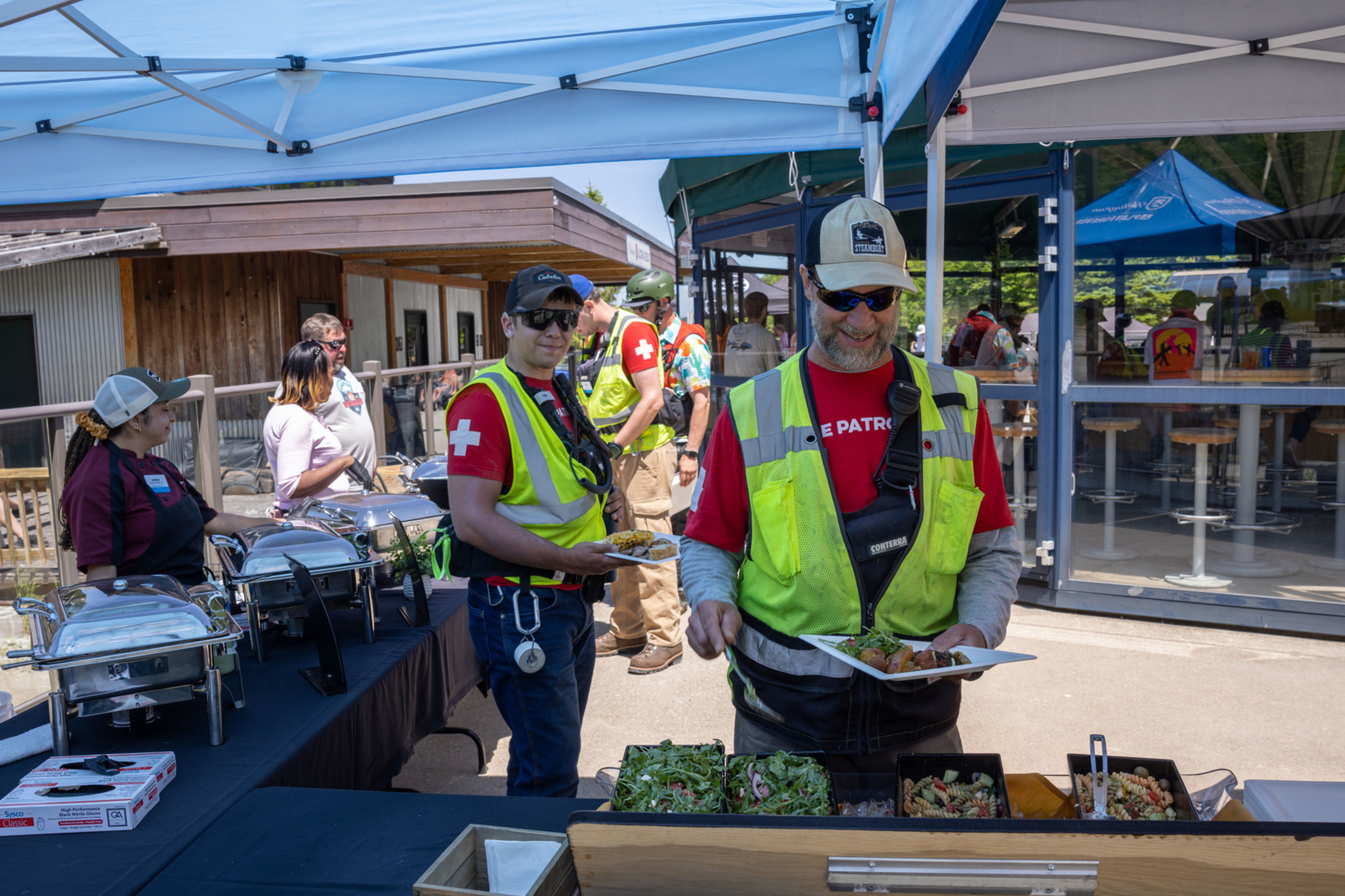 Group of people enjoying a buffet outdoors, wearing safety gear and serving food.
