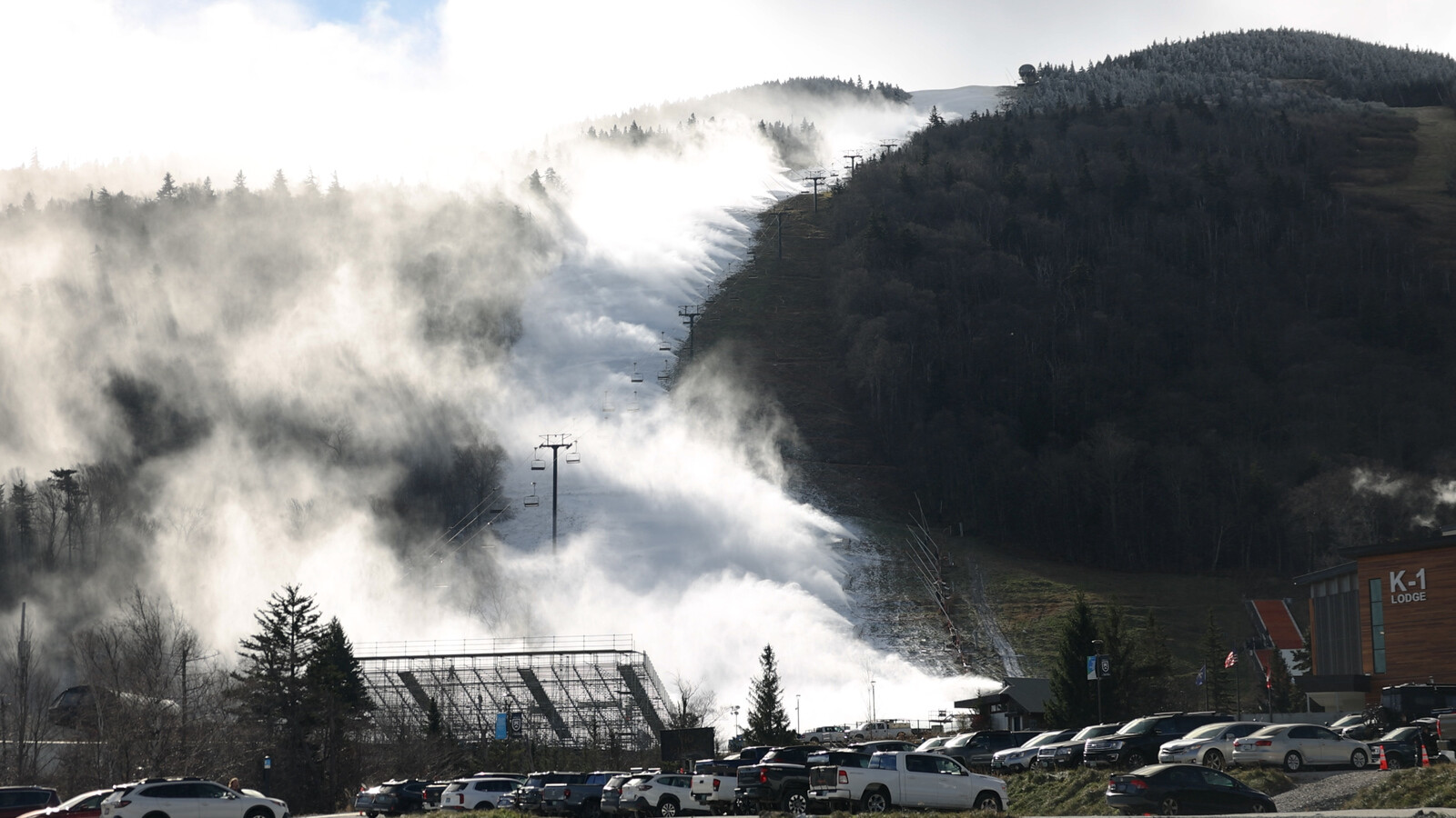 Ski resort with mist and snowmaking equipment on the slopes, parking lot in the foreground.