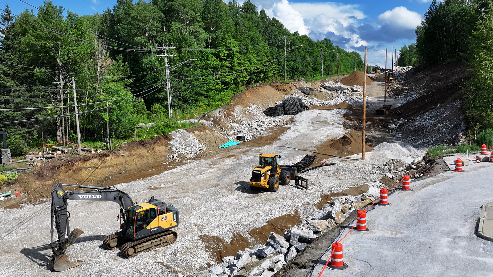 Construction site with heavy machinery, gravel, and electric poles surrounded by trees.