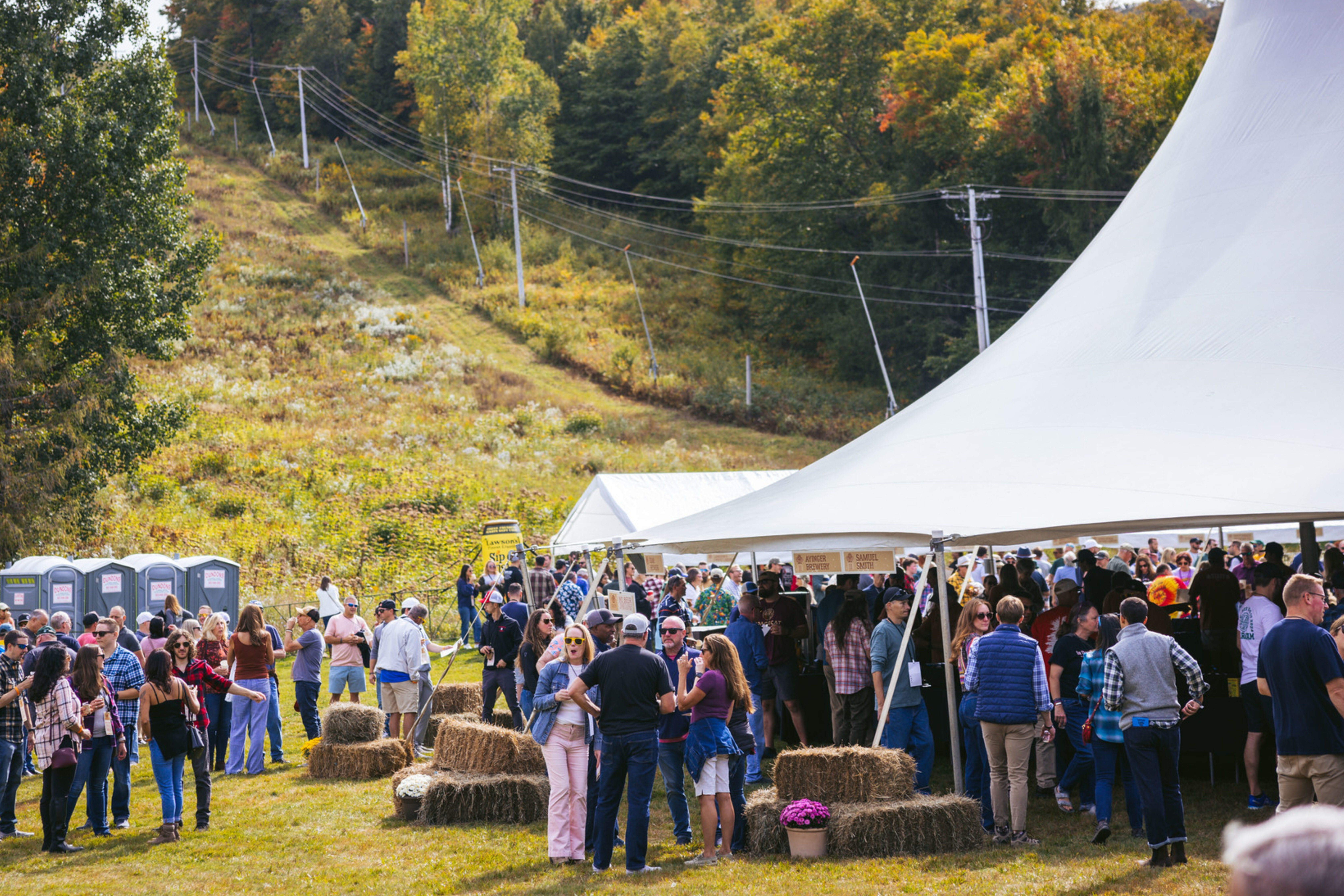 A crowded outdoor festival with people, tents, and autumn foliage in the background.