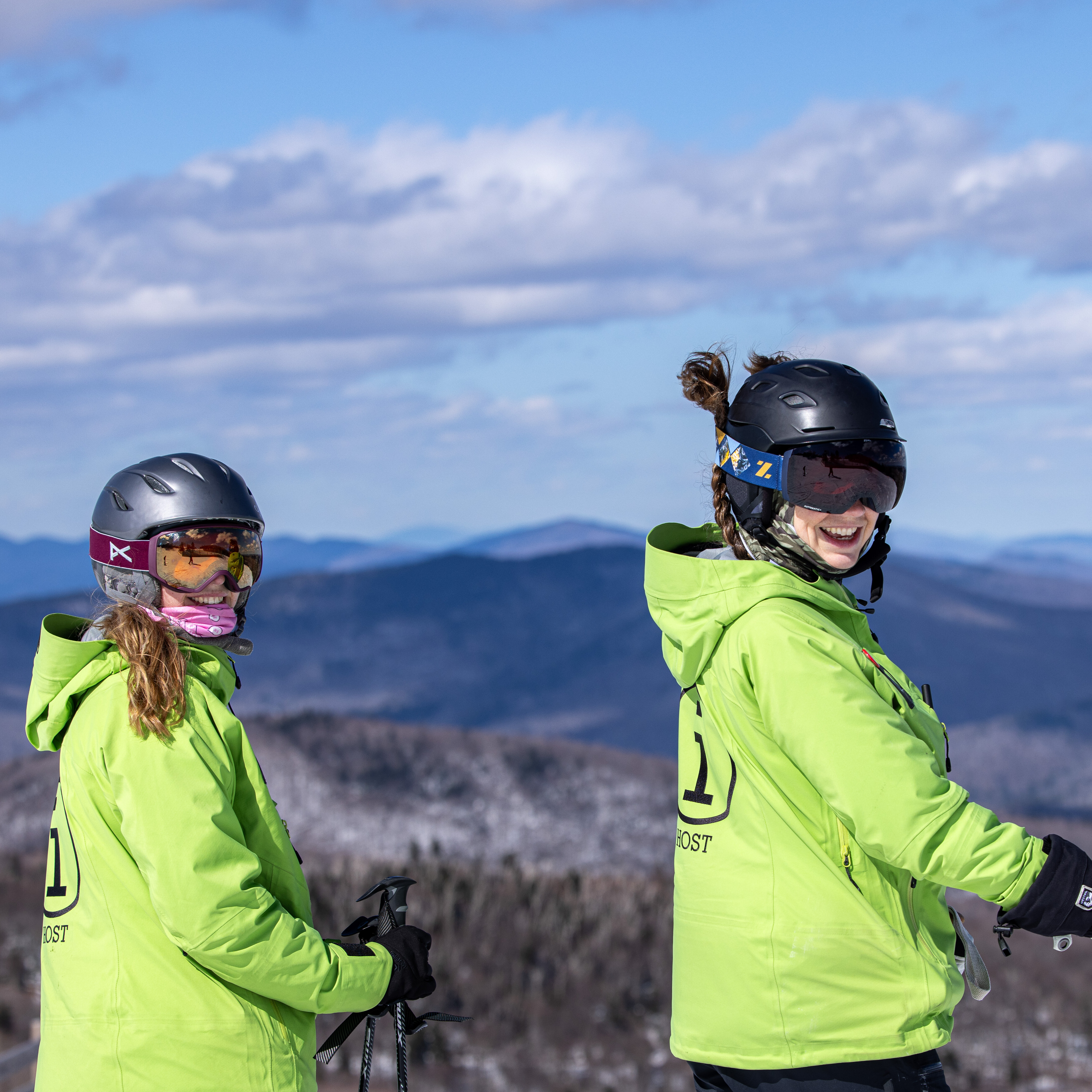 Two skiers in bright green jackets smile against a mountainous backdrop.