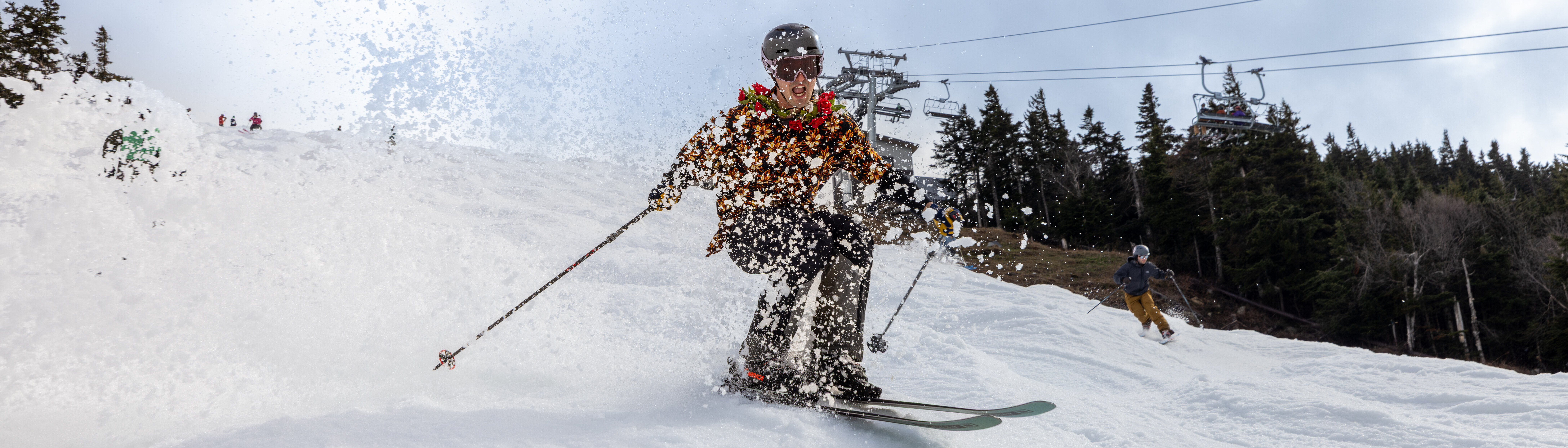 A skier in a colorful jacket sprays snow while skiing down a slope, with trees and a chairlift in the background.