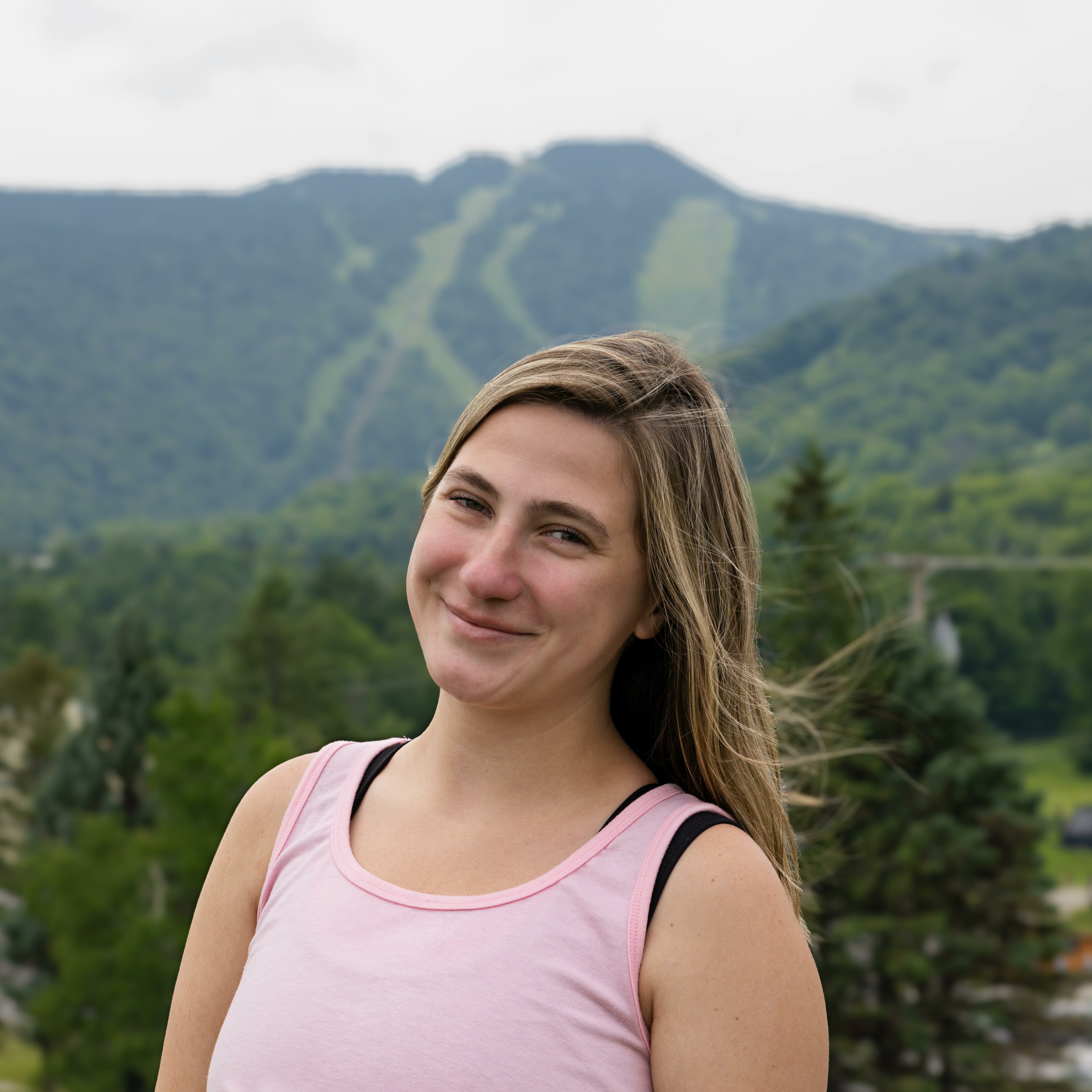 A smiling young woman stands outdoors with a mountain landscape in the background.