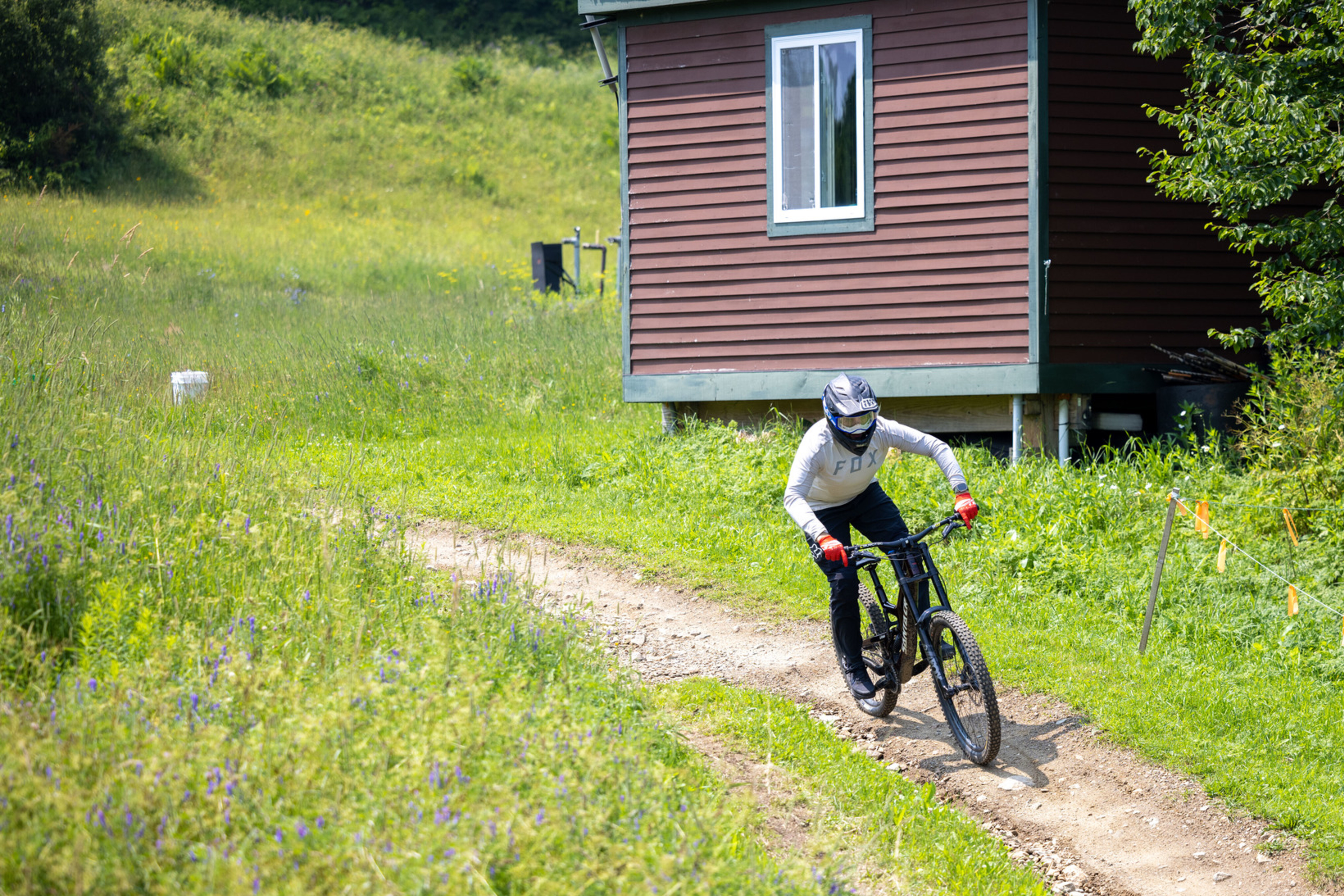 A mountain biker rides on a dirt path near a small brown house in a grassy area.