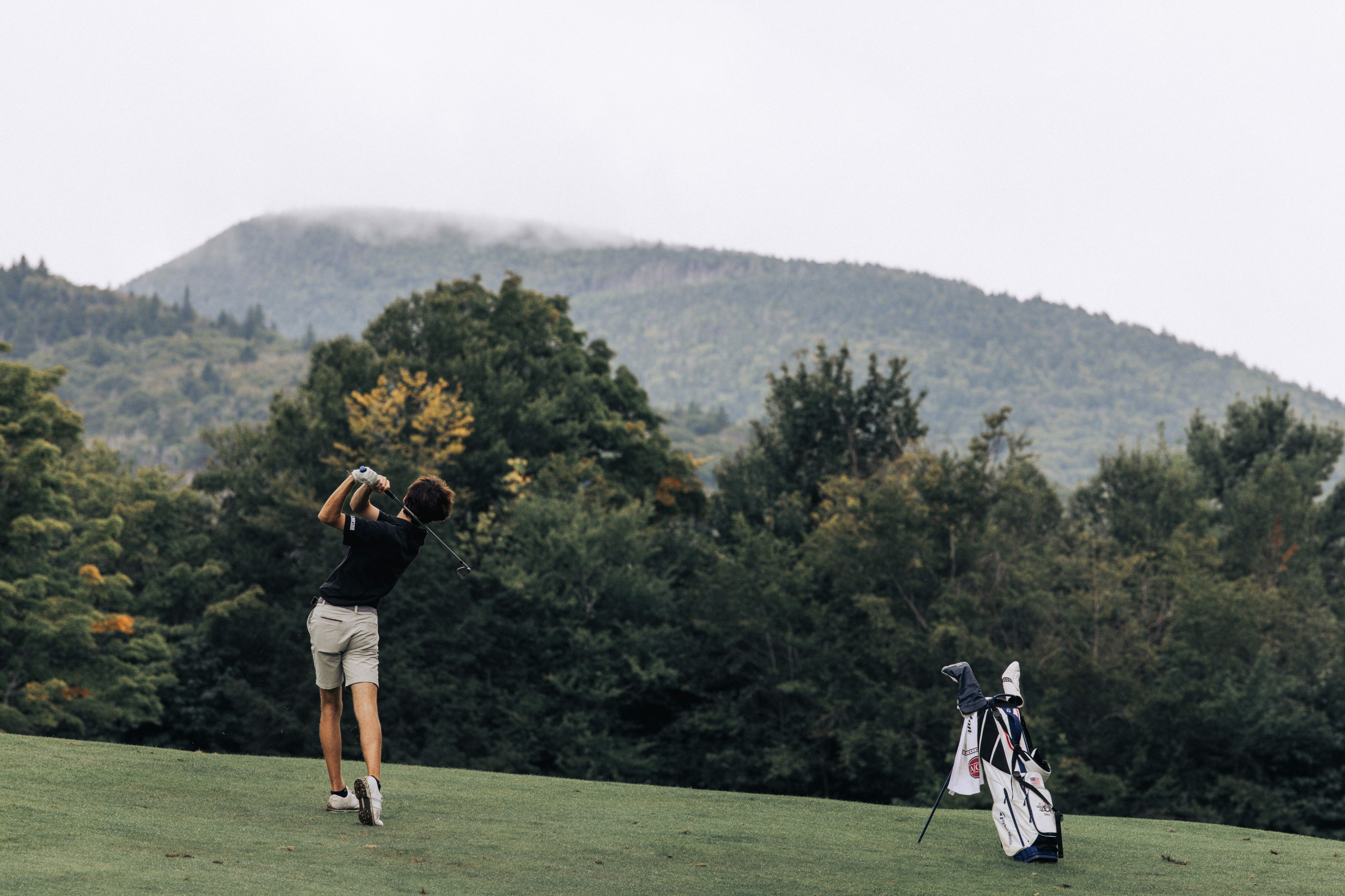 A golfer swings on a course with mountains and trees in the background.