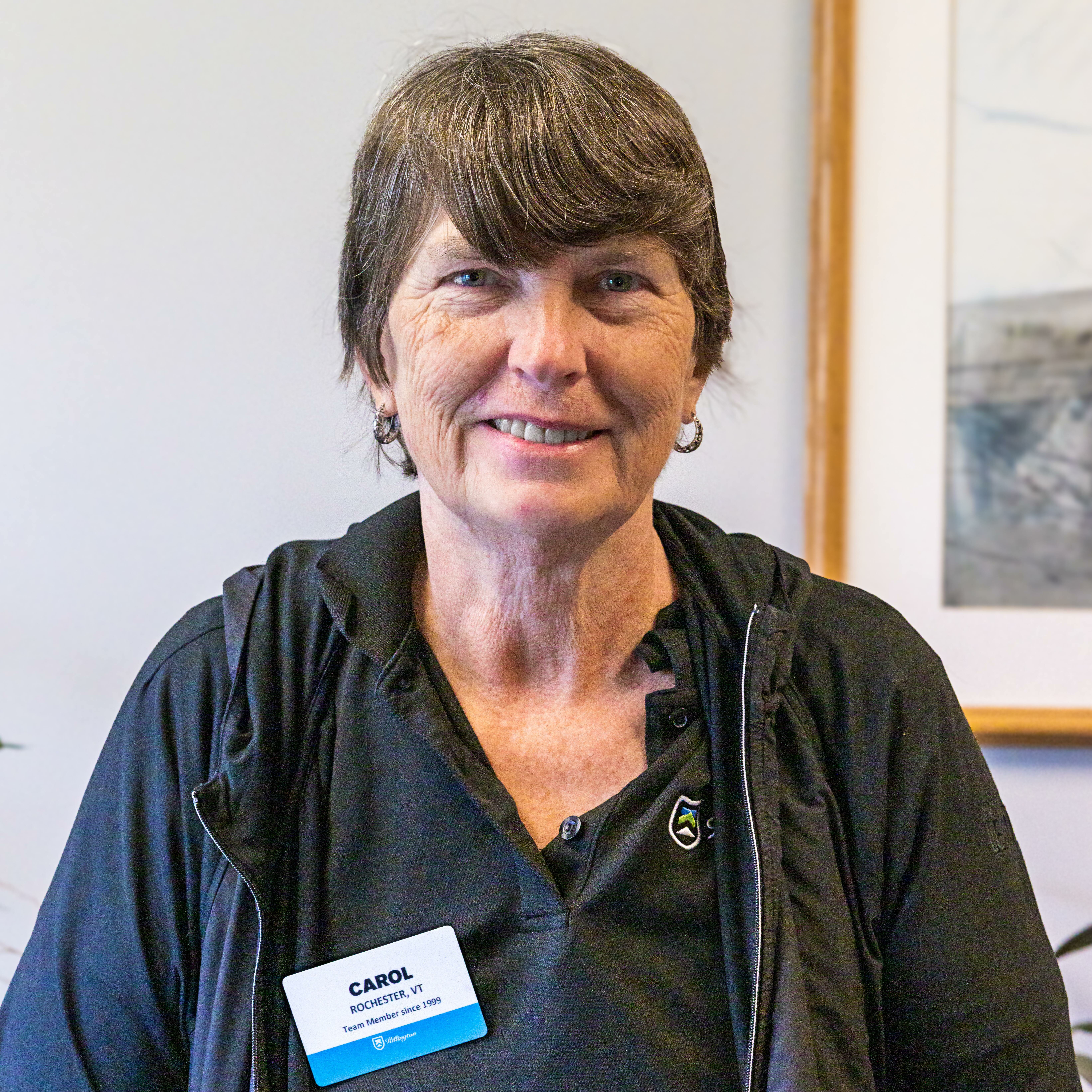 Portrait of a woman named Carol, wearing a name tag and smiling at the camera.