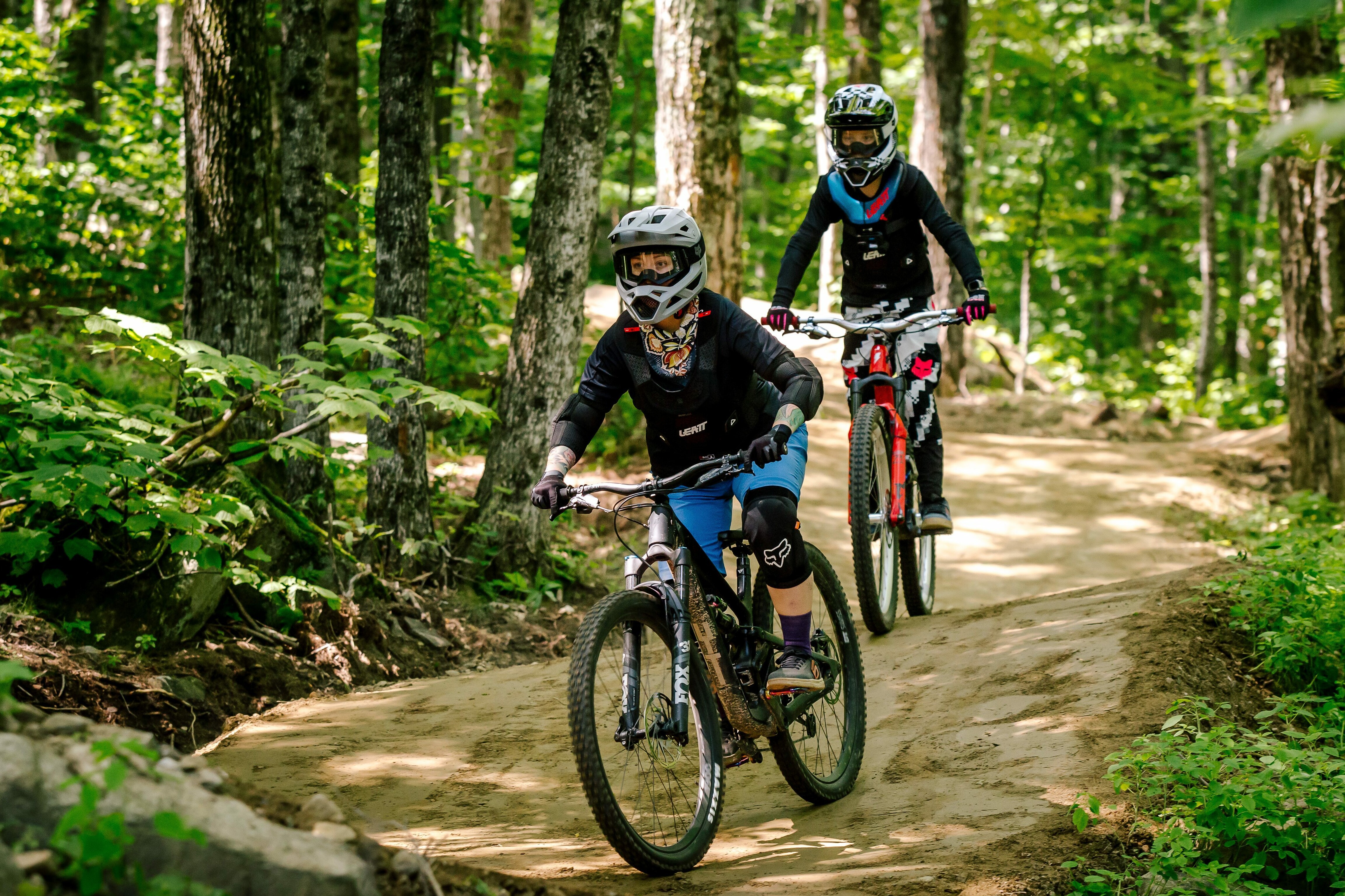 Two mountain bikers ride on a dirt trail surrounded by green trees.