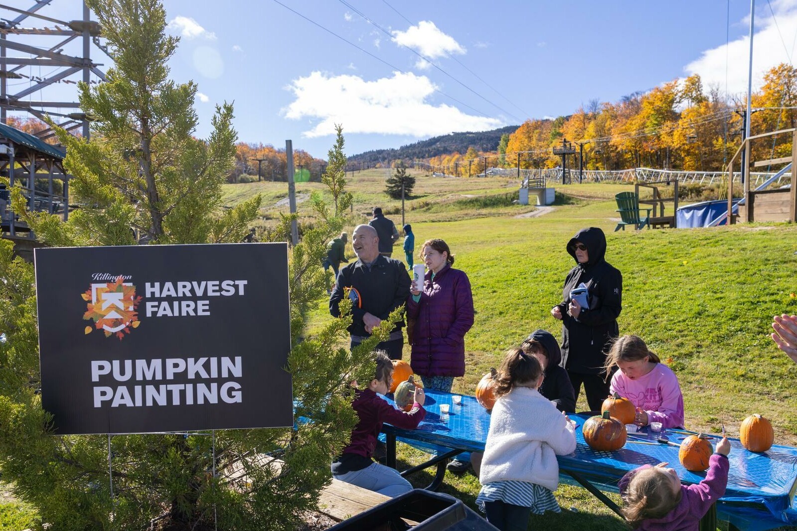 Families enjoy pumpkin painting at the Harvest Faire, with autumn scenery in the background.