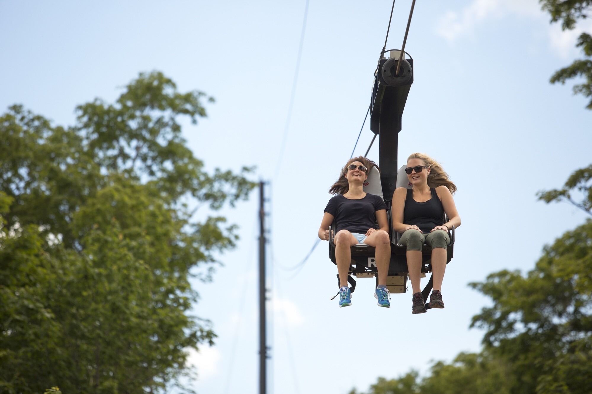 Two women smile as they ride a chairlift against a blue sky and green trees.
