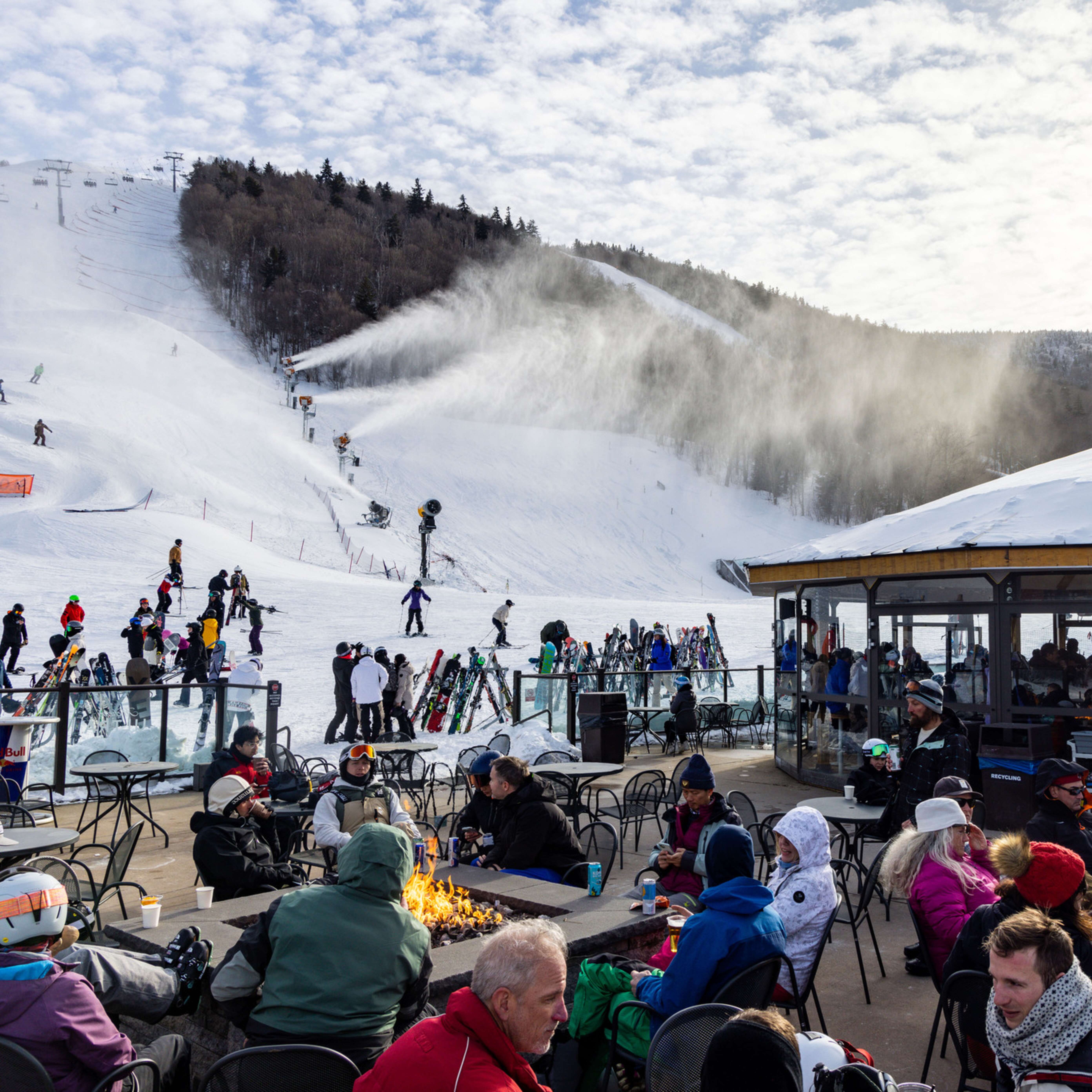 Ski resort scene with people enjoying the outdoors, snowmaking machines, and a cozy fire pit.