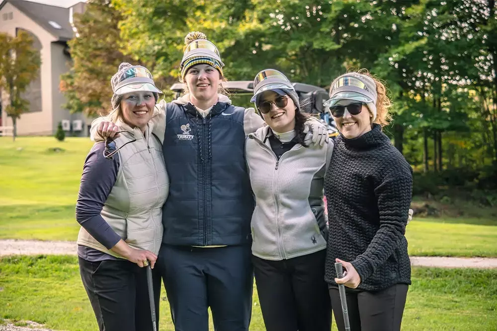 Four women pose together on a golf course, smiling and holding golf clubs.