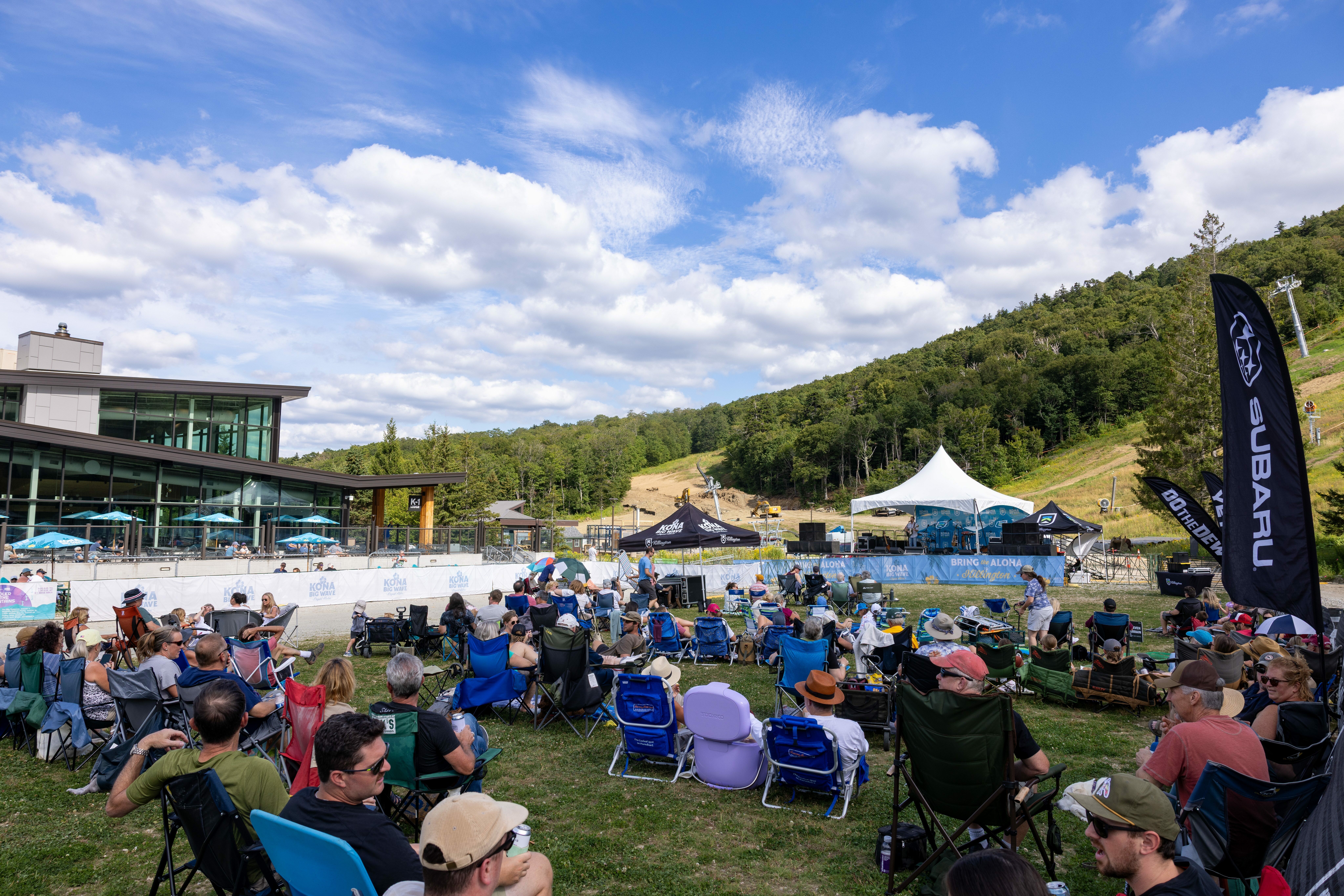 Outdoor concert scene with crowd in chairs, mountain backdrop, and stage.