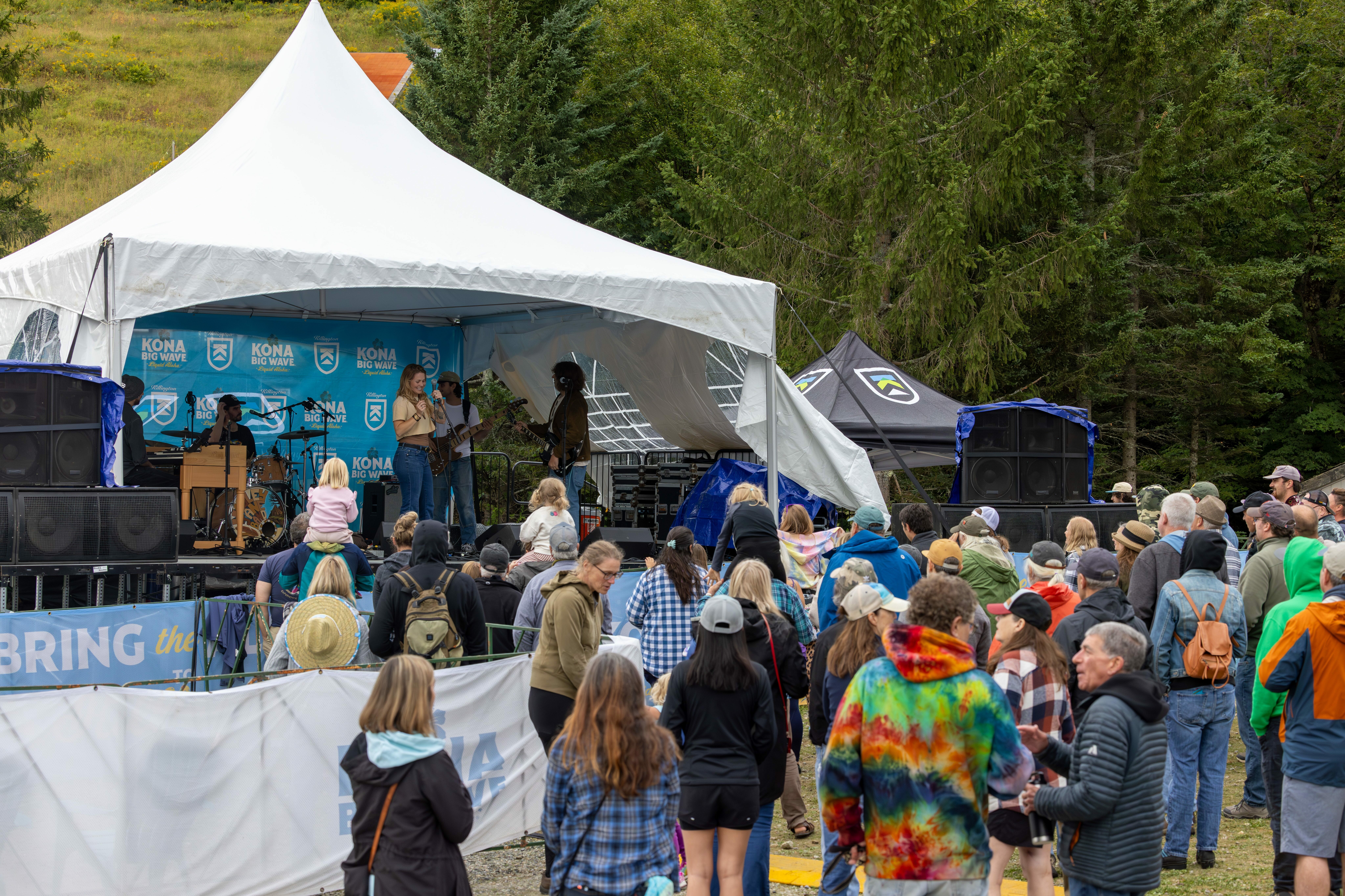 A crowd enjoys live music at an outdoor festival stage under a large tent.