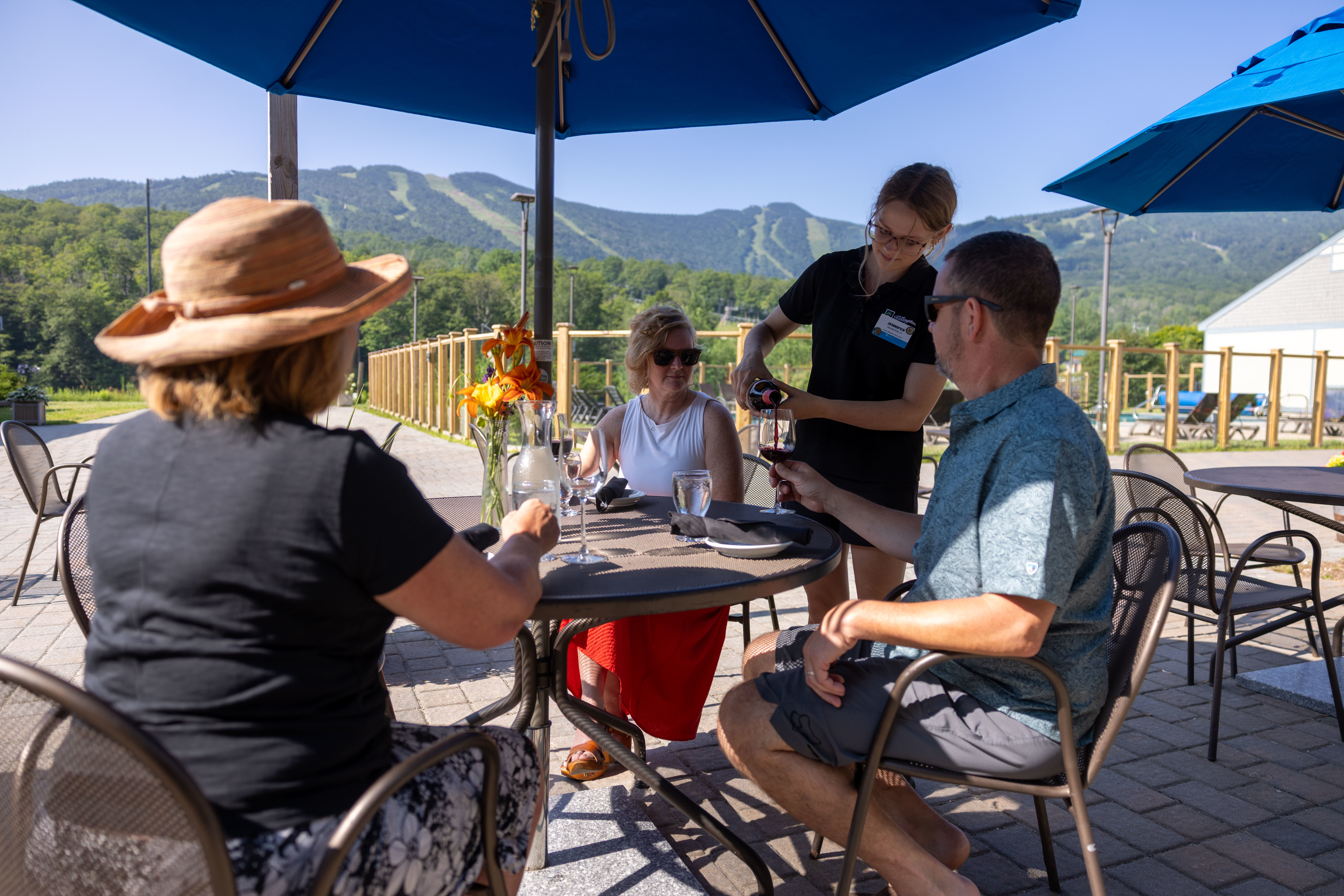 Four people dining outdoors at a table with a mountain view, a server pouring wine.