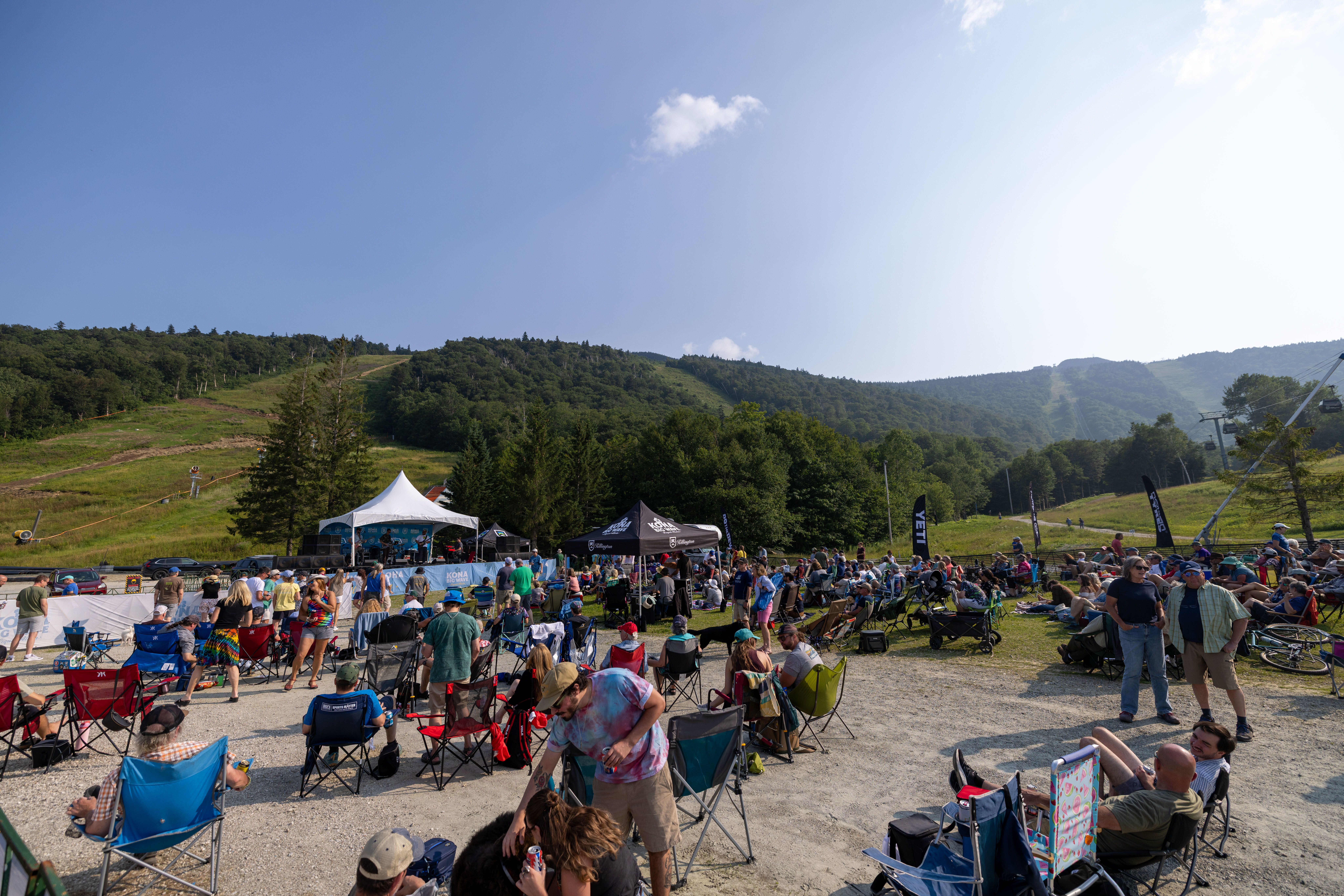 Outdoor concert crowd with chairs in front of a stage, surrounded by mountains and trees.