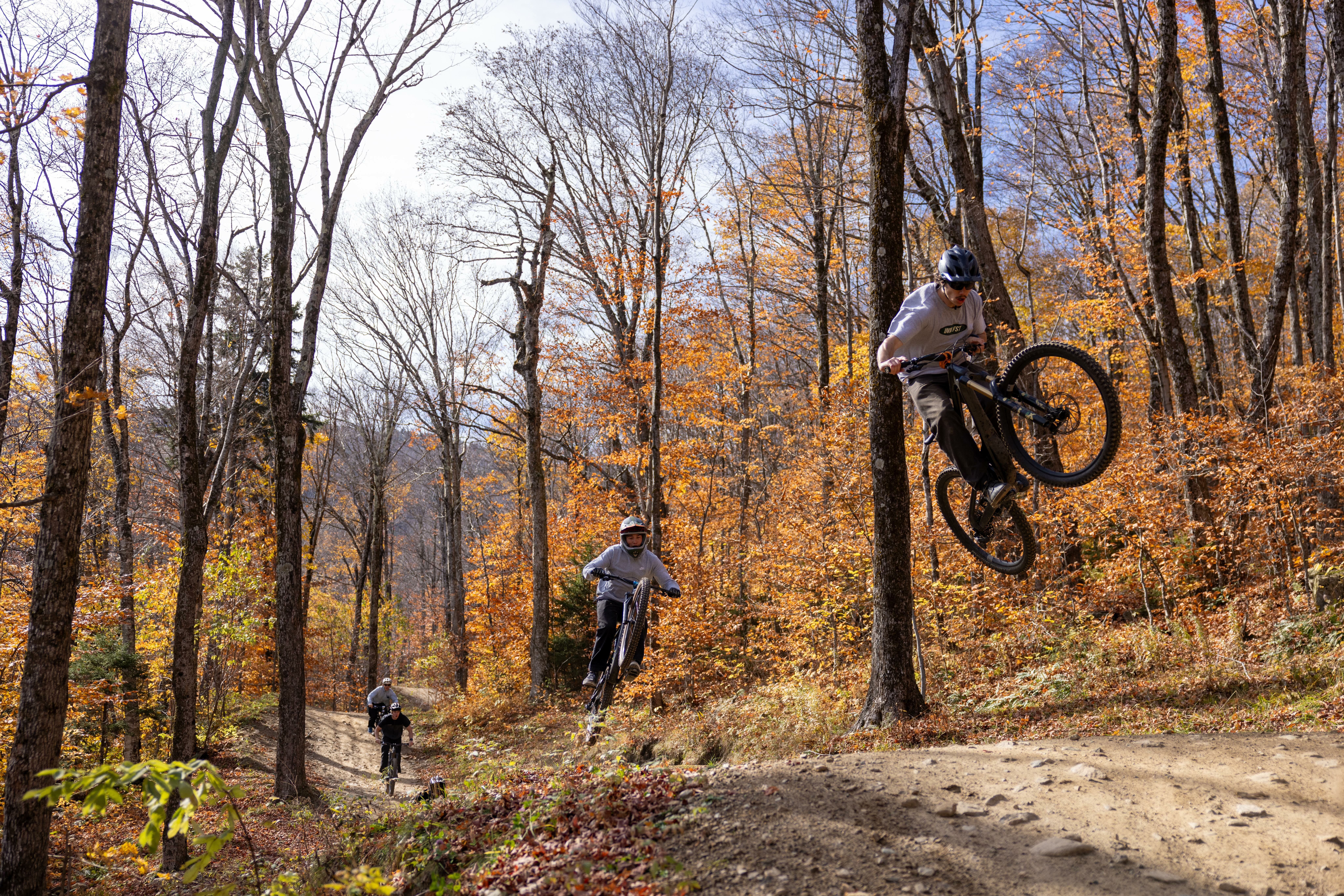 Two mountain bikers ride on a dirt path through vibrant autumn trees.