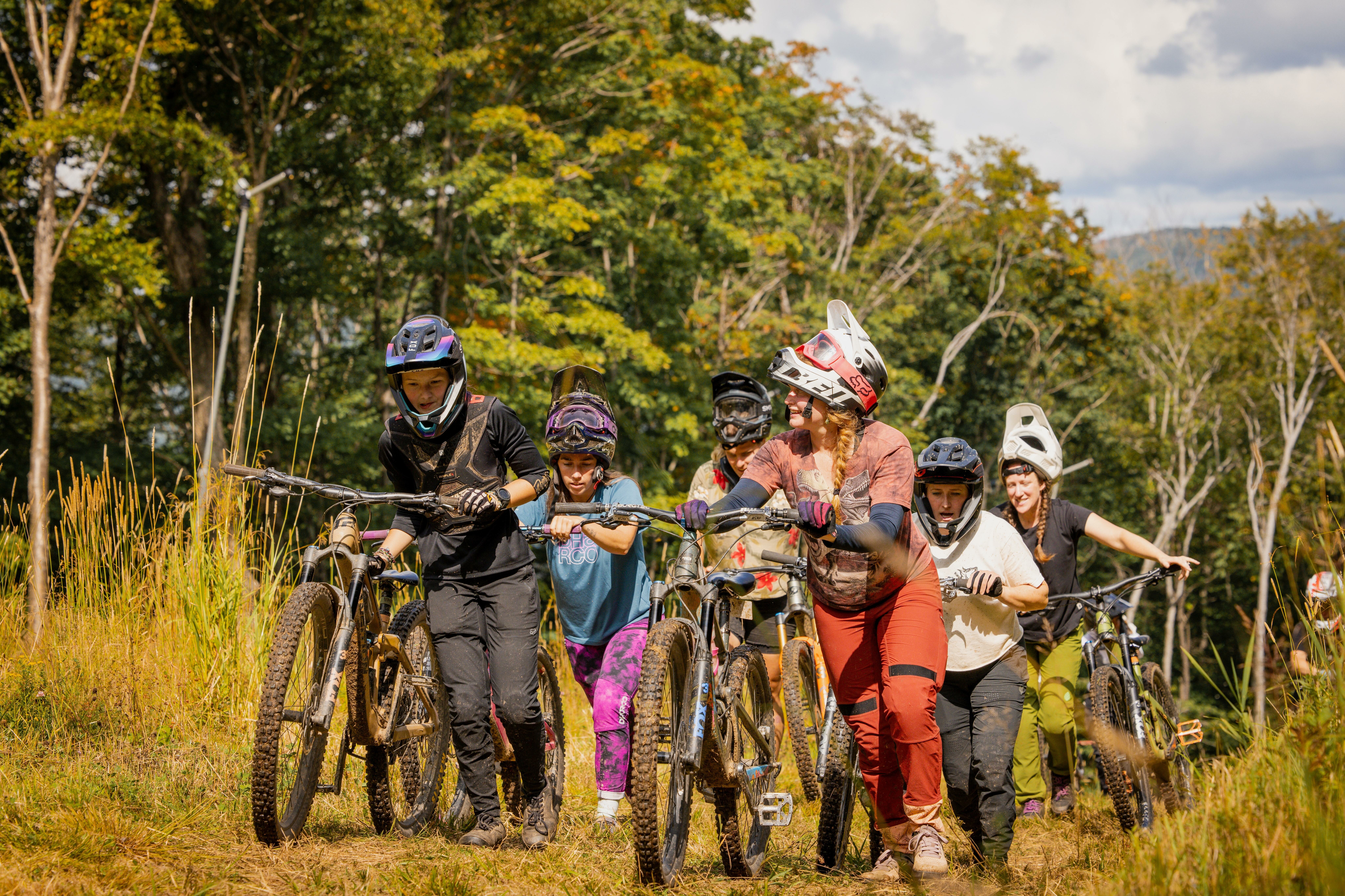 A group of young bikers pushing their mountain bikes uphill in a lush, green landscape.