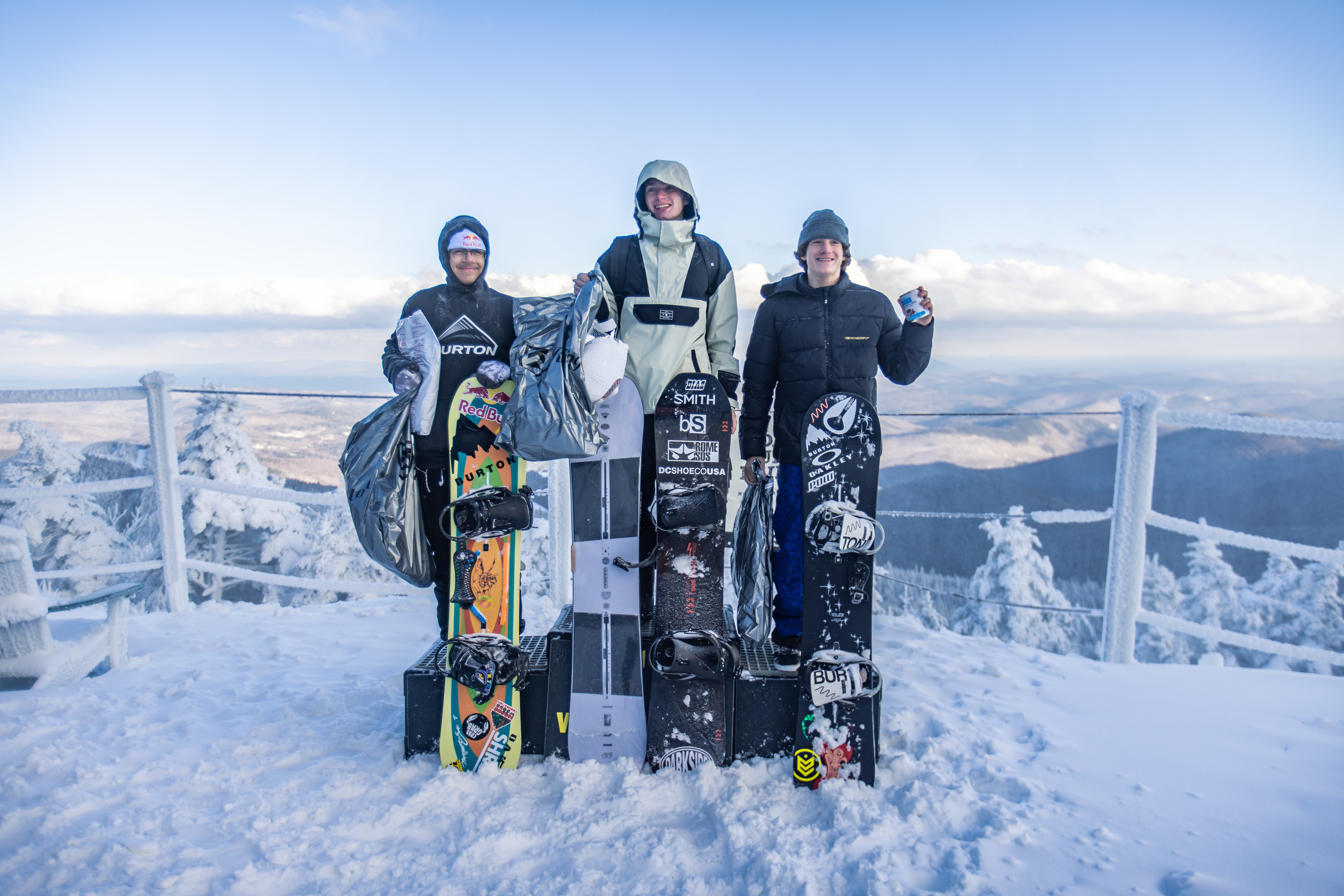 Three snowboarders pose on a snowy mountain peak with their boards and gear.