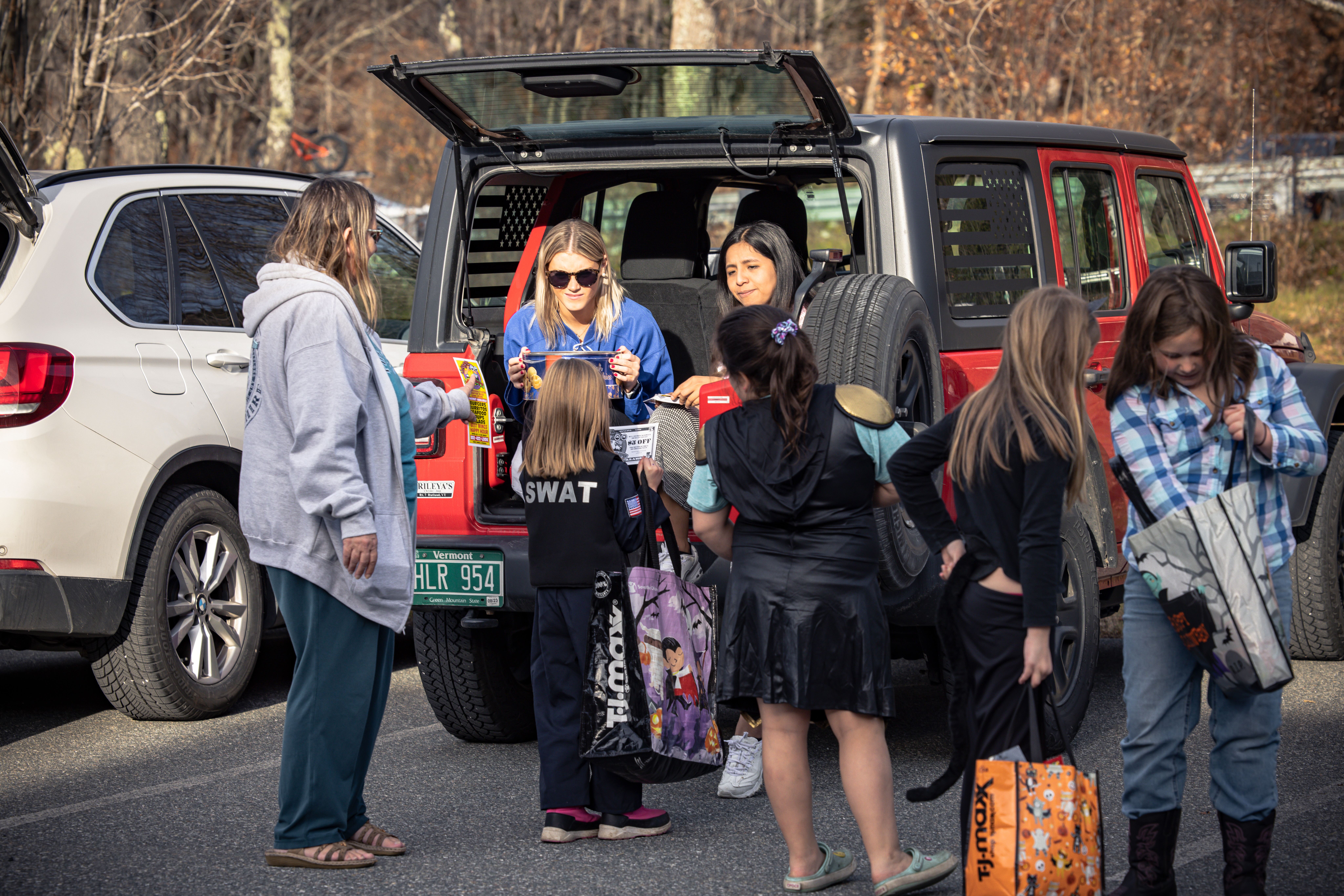A group of children engage with two women at a car, sharing treats and Halloween bags.