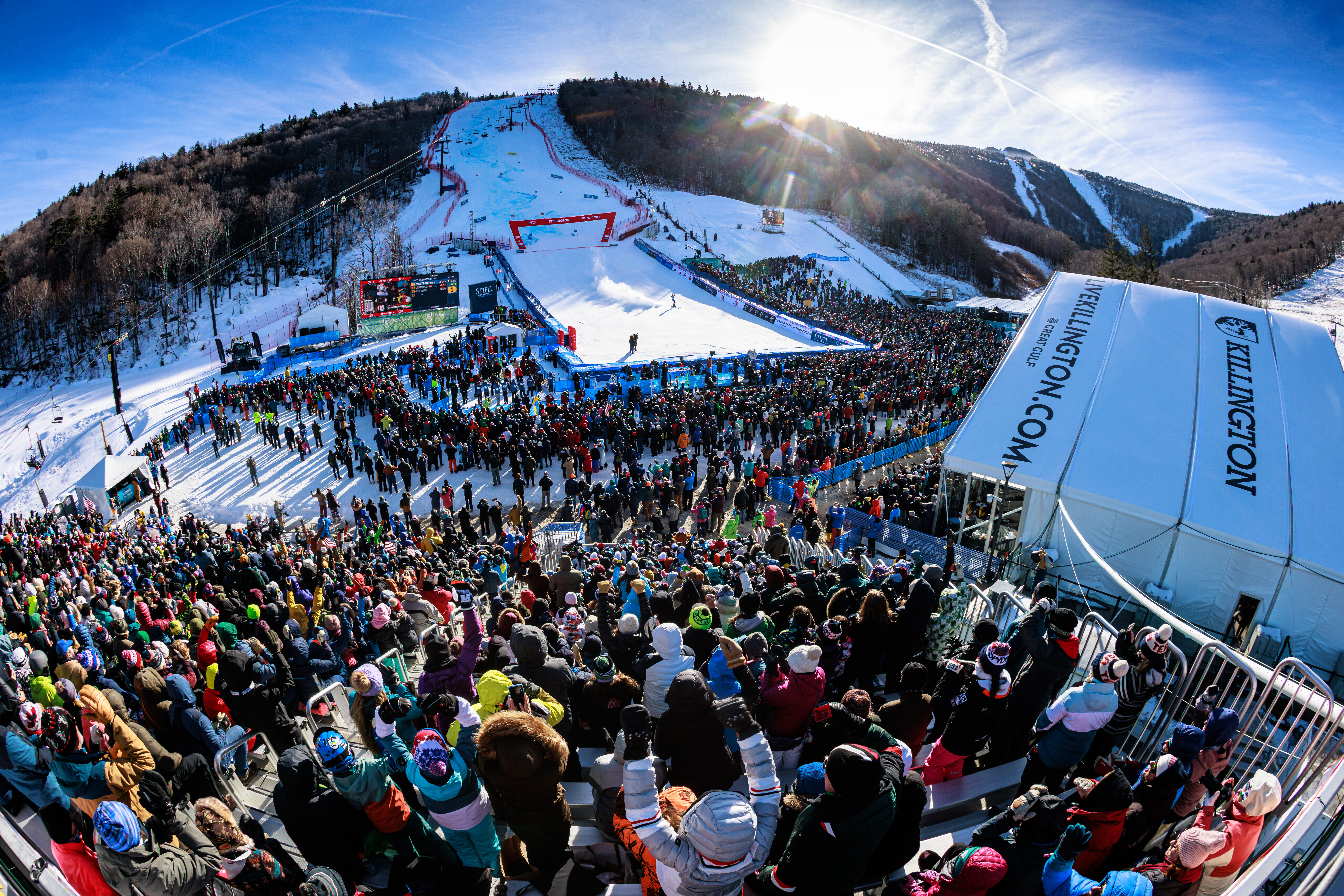 Crowd gathered at a snowy ski resort for a winter sports event under clear blue skies.