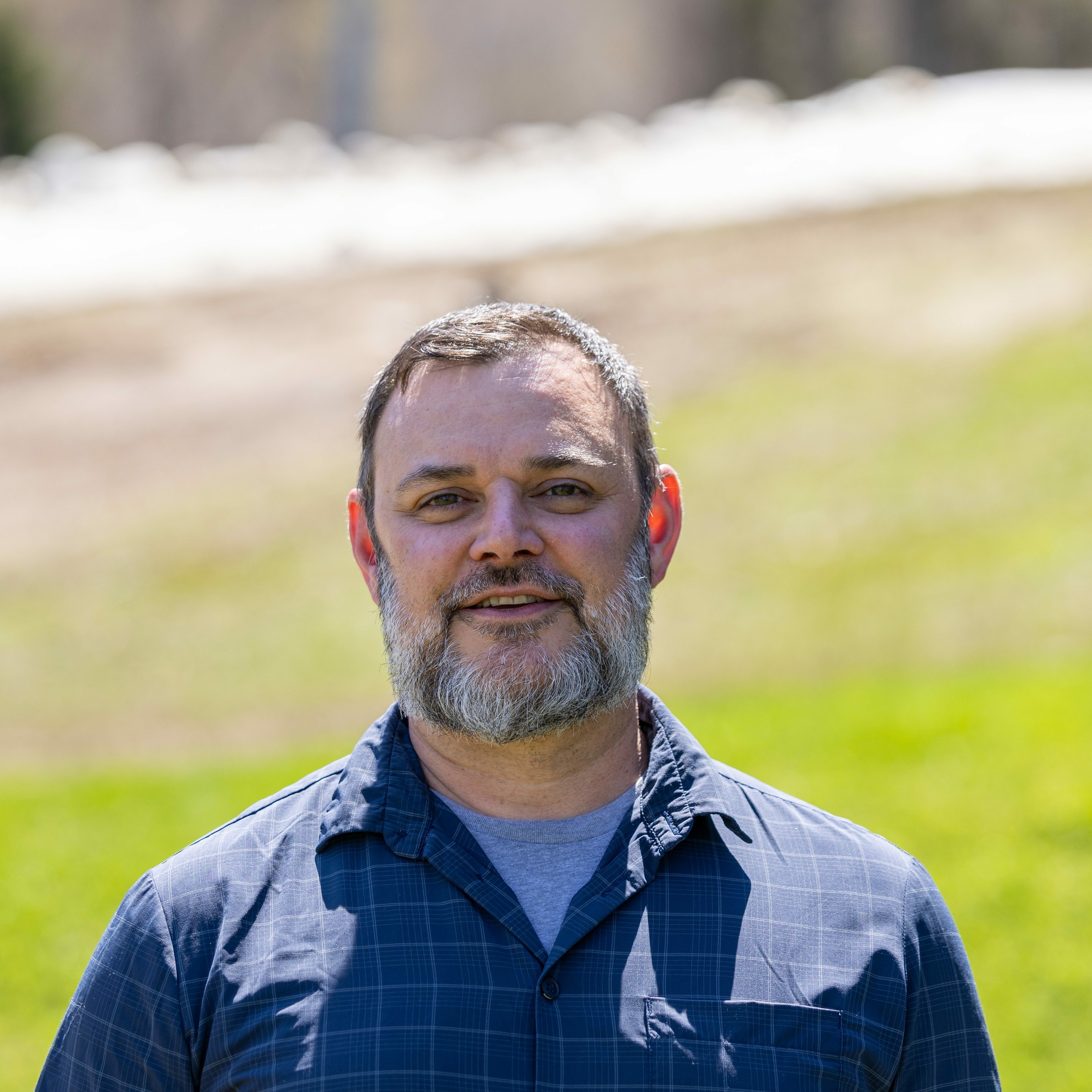 A smiling man with a beard stands outdoors in front of a green field.