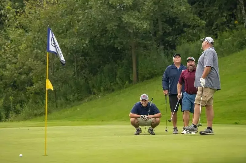 Four men on a golf course, discussing a shot near the hole with a flag in the background.