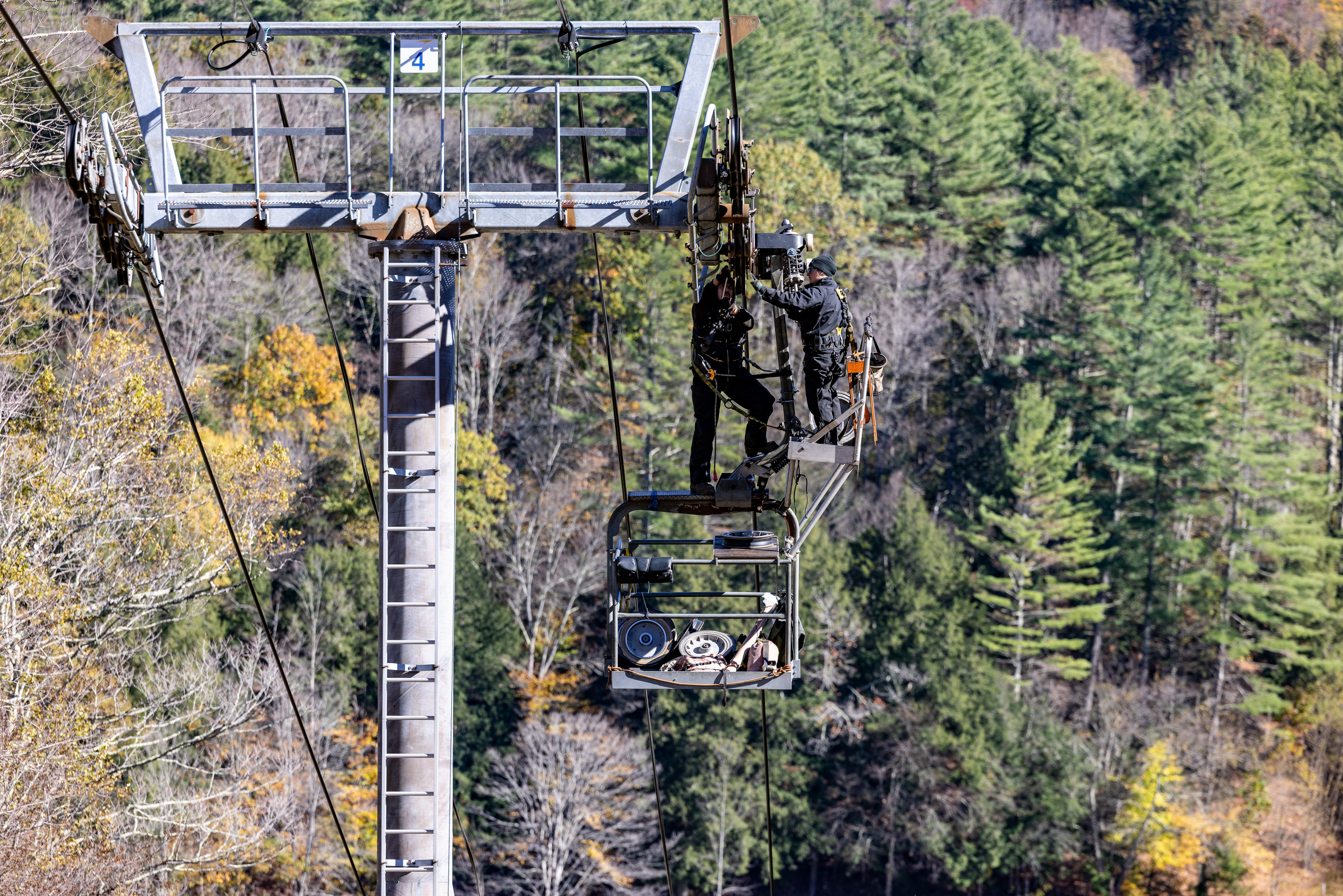 Two workers perform maintenance on a gondola high above forested trees.