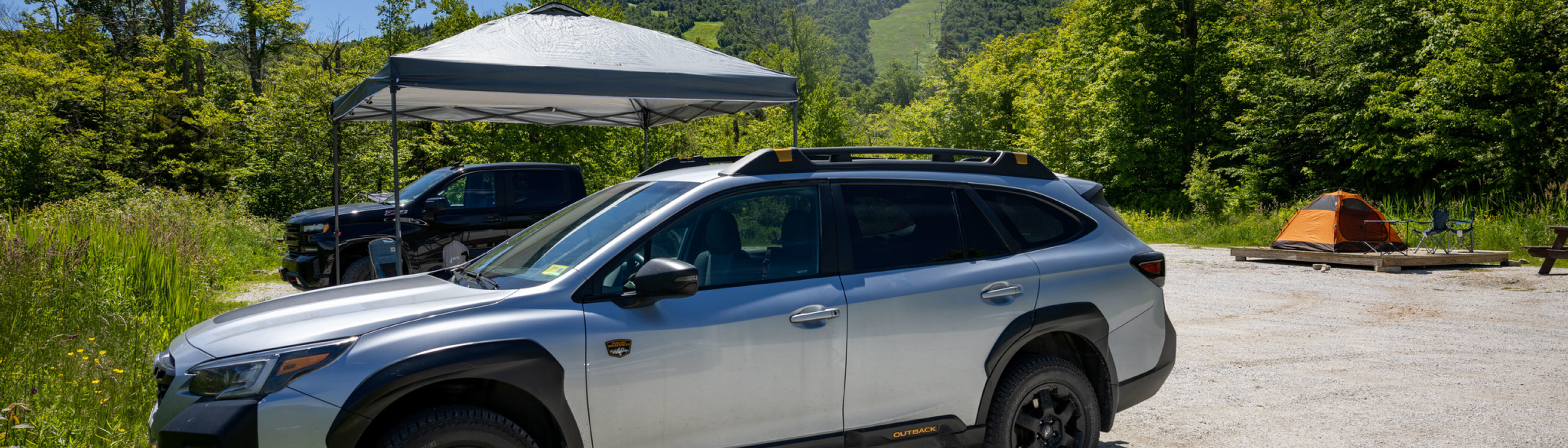 A silver SUV parked near a tent and camping chairs in a green outdoor setting.