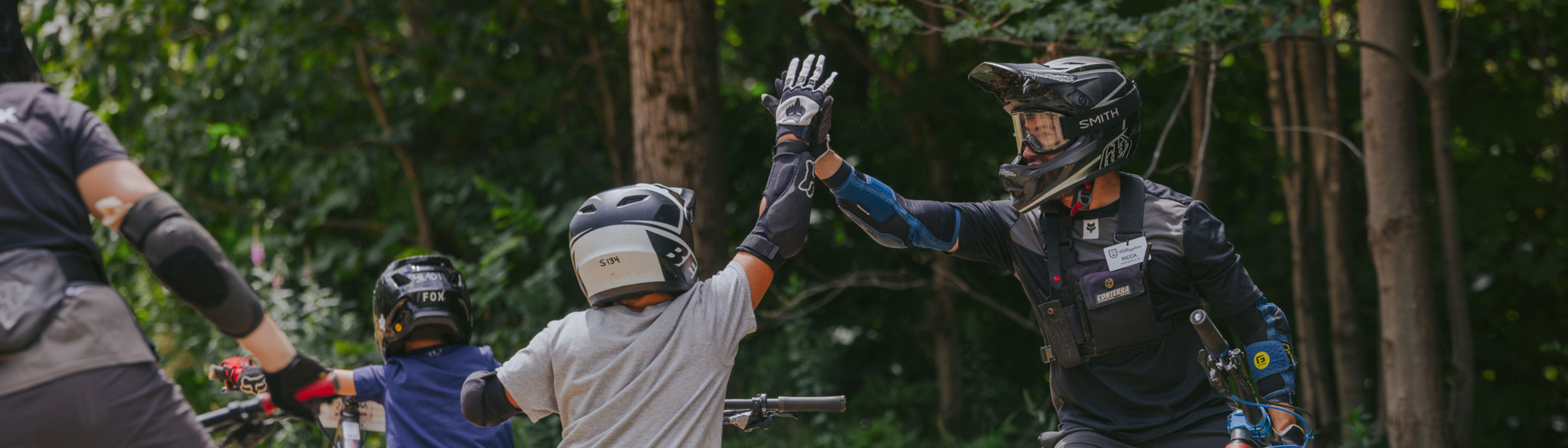 A helmeted instructor high-fives a child on a bike, both in protective gear, outdoors.
