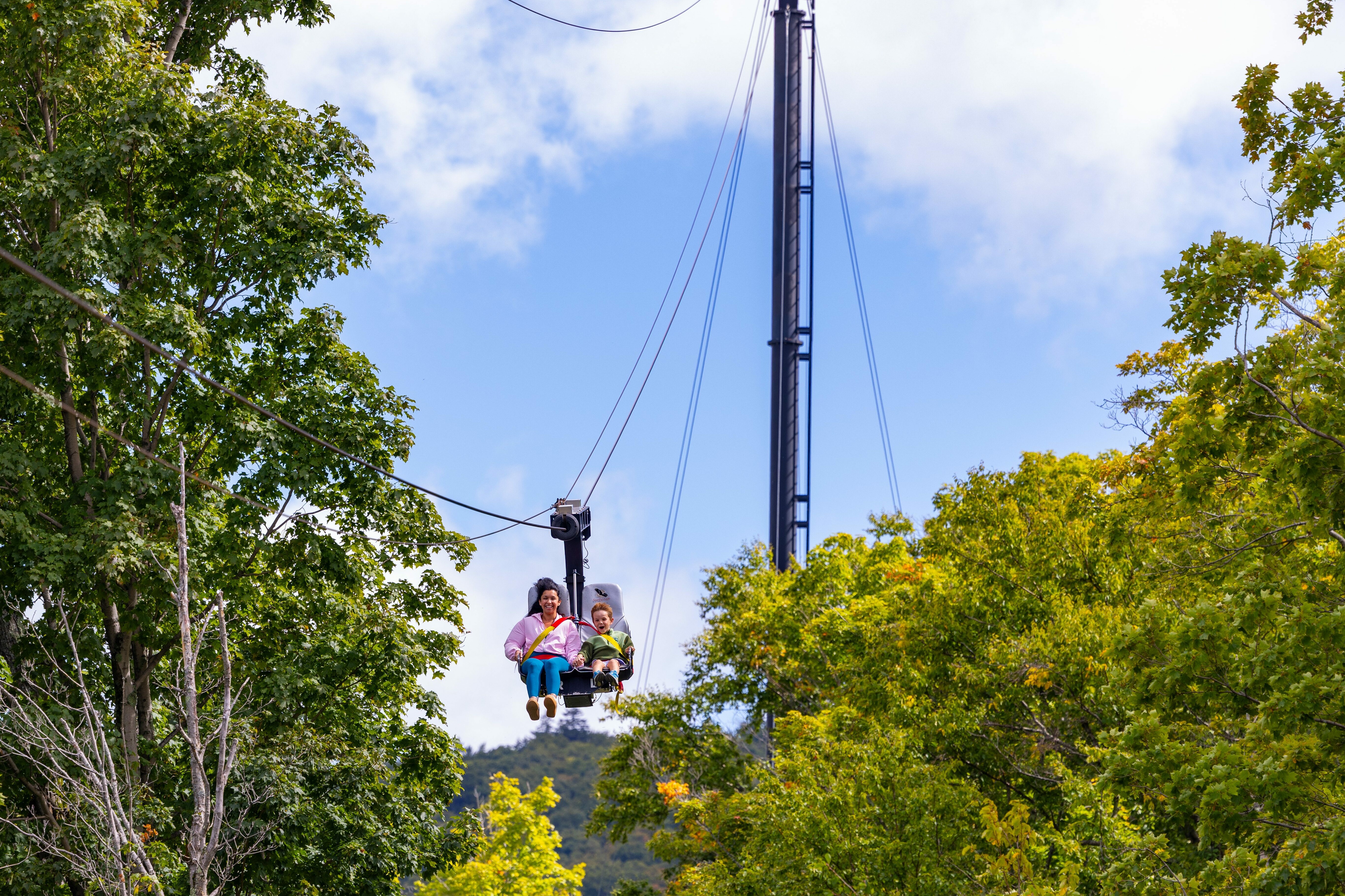 A mother and child on a chairlift surrounded by trees and a blue sky.