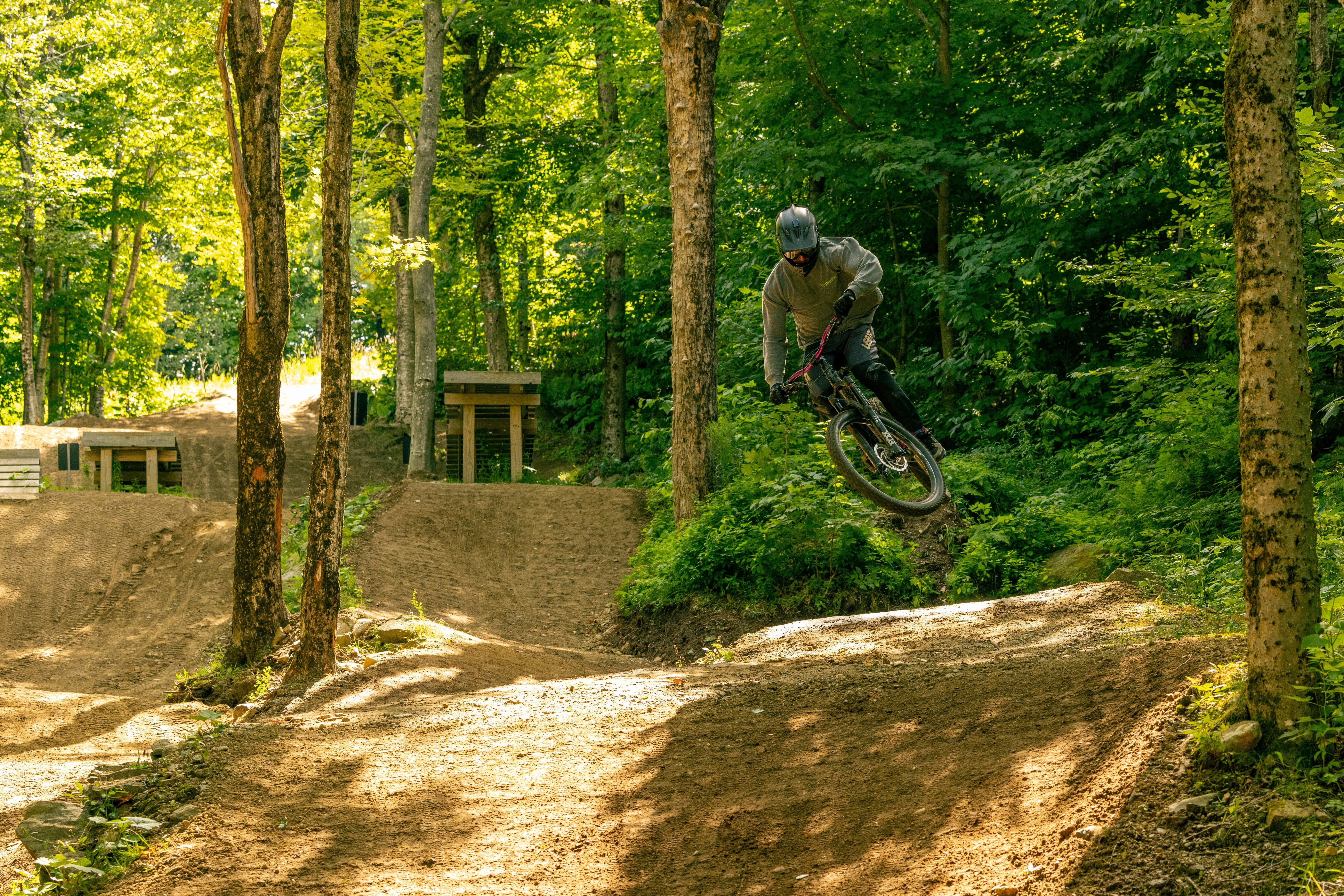 A cyclist performs a jump on a dirt bike trail surrounded by trees.