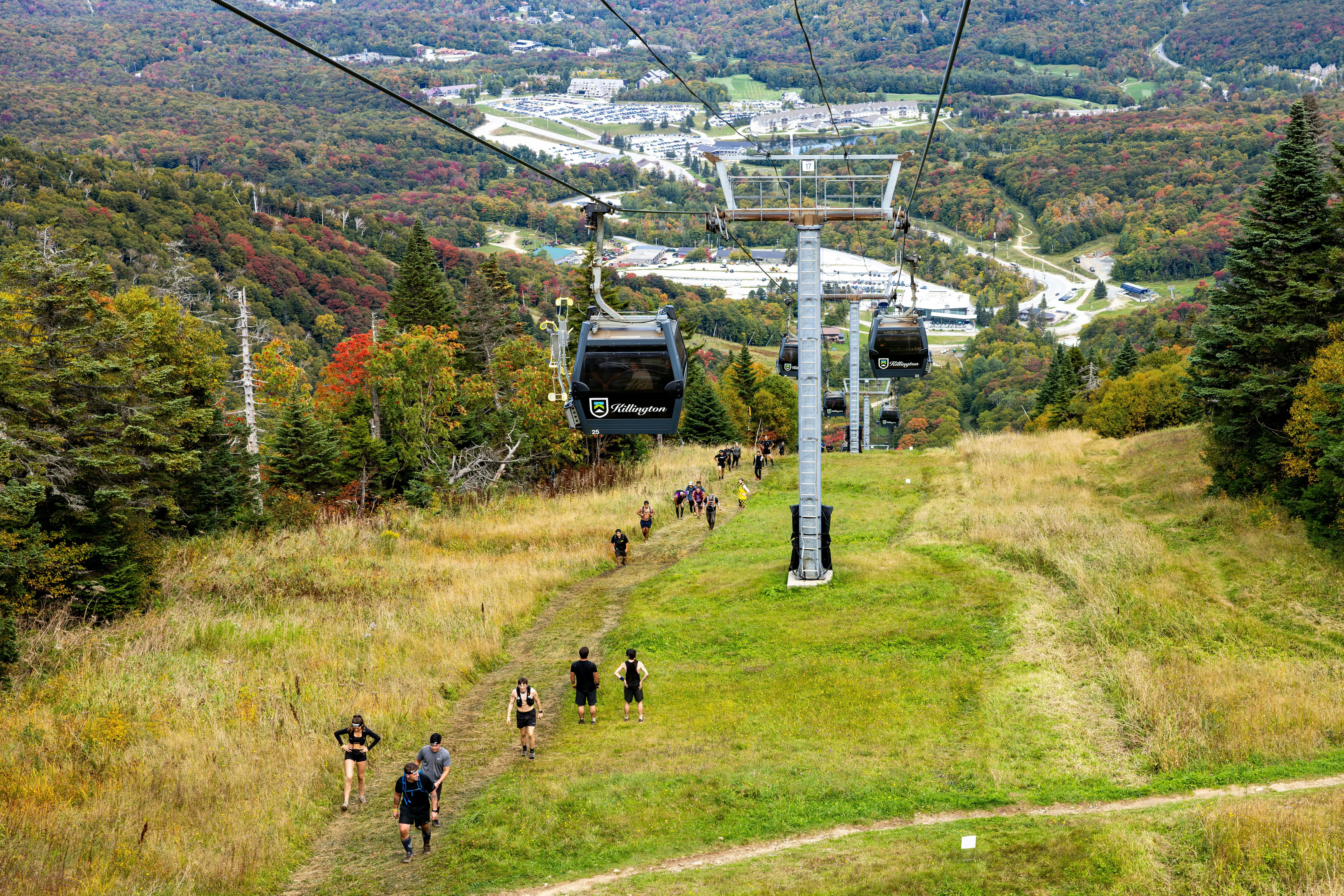 People hike up a grassy slope with a ski lift above and autumn foliage in the background.
