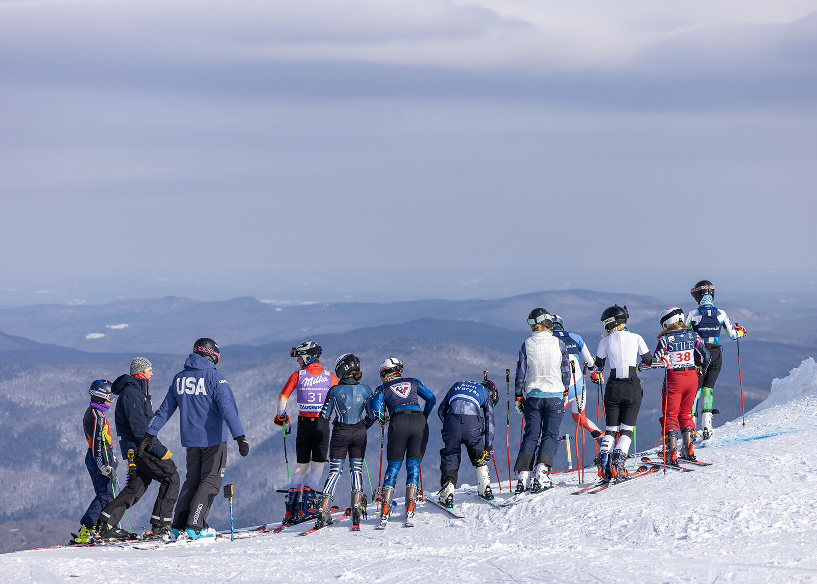 Skiers prepare at the edge of a snowy slope, overlooking distant mountains.