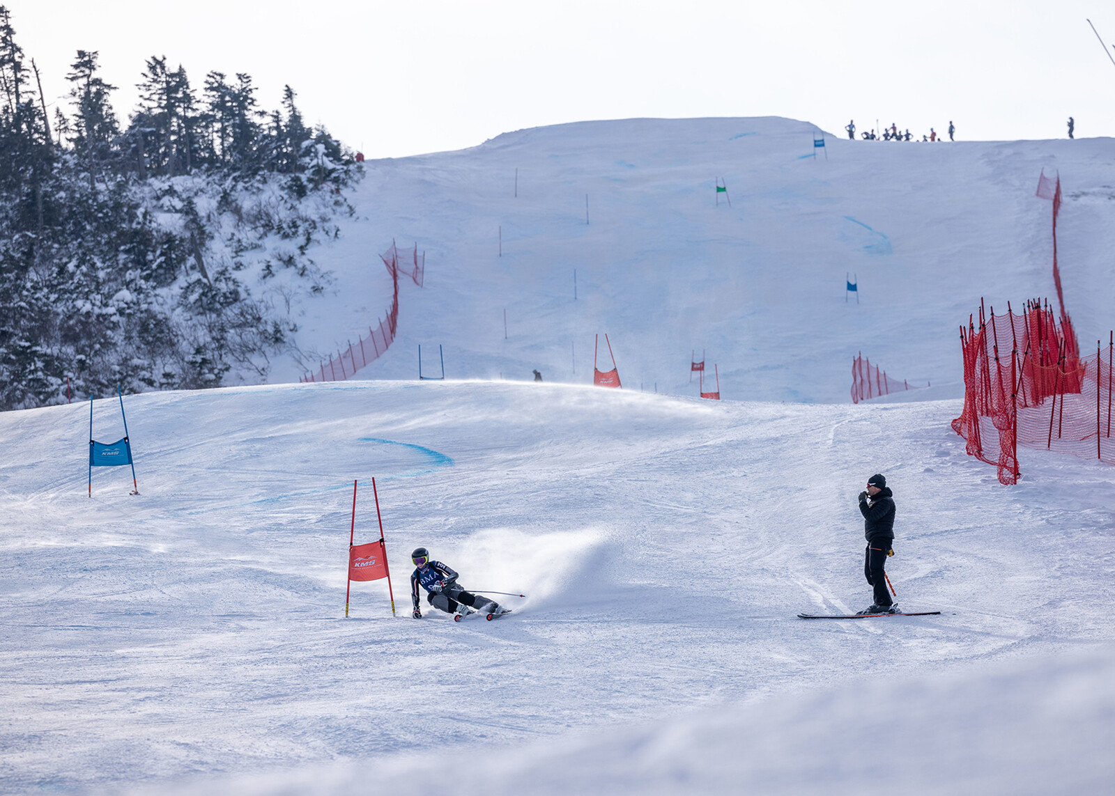A skier navigates a snowy course marked by gates while another person watches nearby.