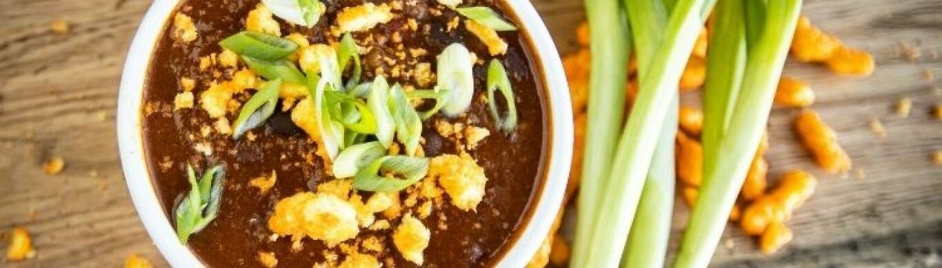 Bowl of dark soup topped with green onions and crispy toppings, beside fresh scallions.