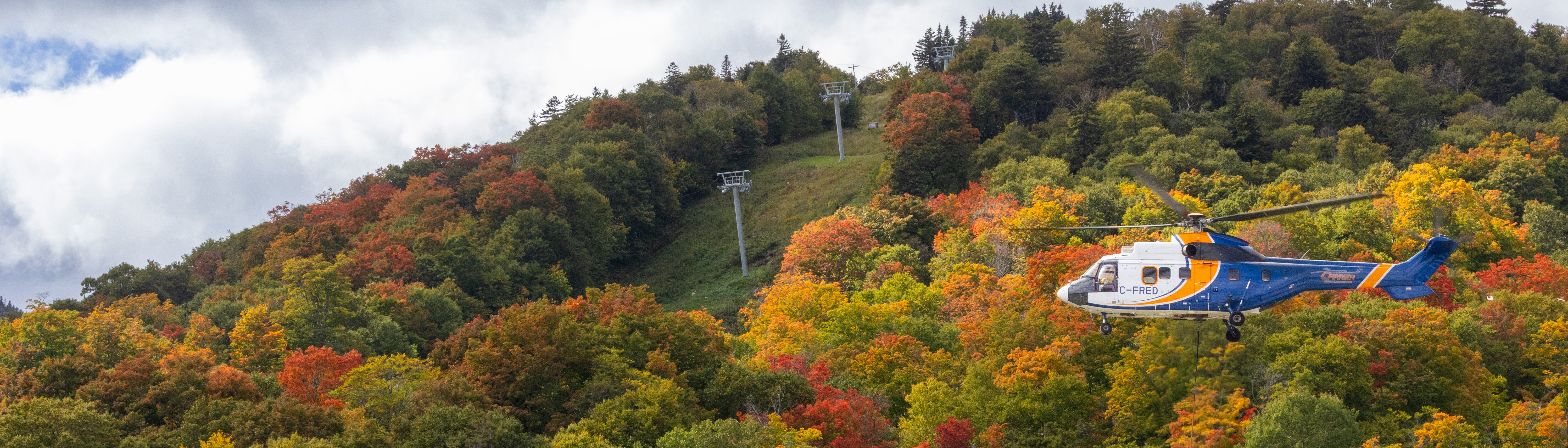 A helicopter flies over a vibrant autumn landscape with colorful foliage and ski lifts.