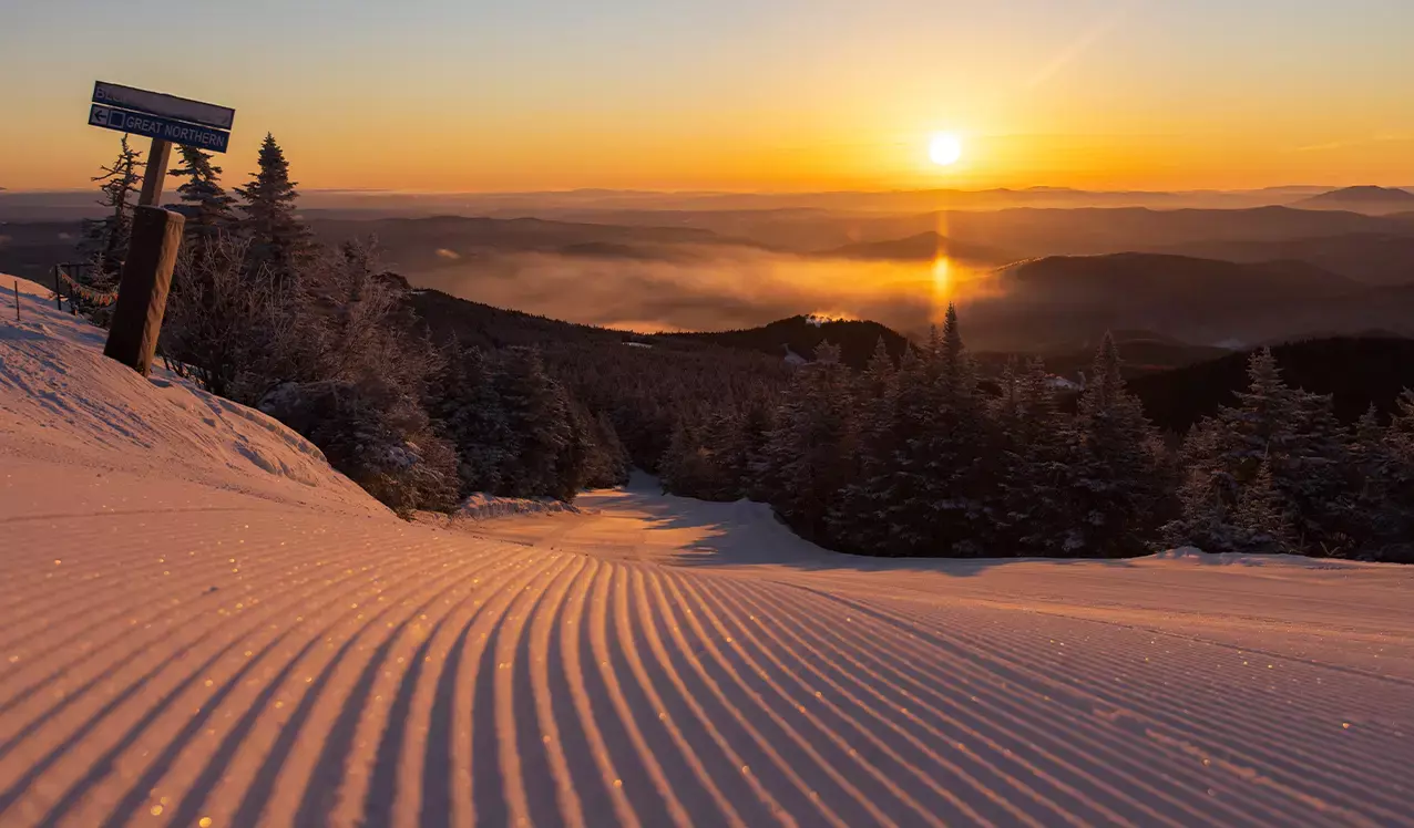 Sunset over snow-covered mountains with groomed ski trails and evergreen trees.