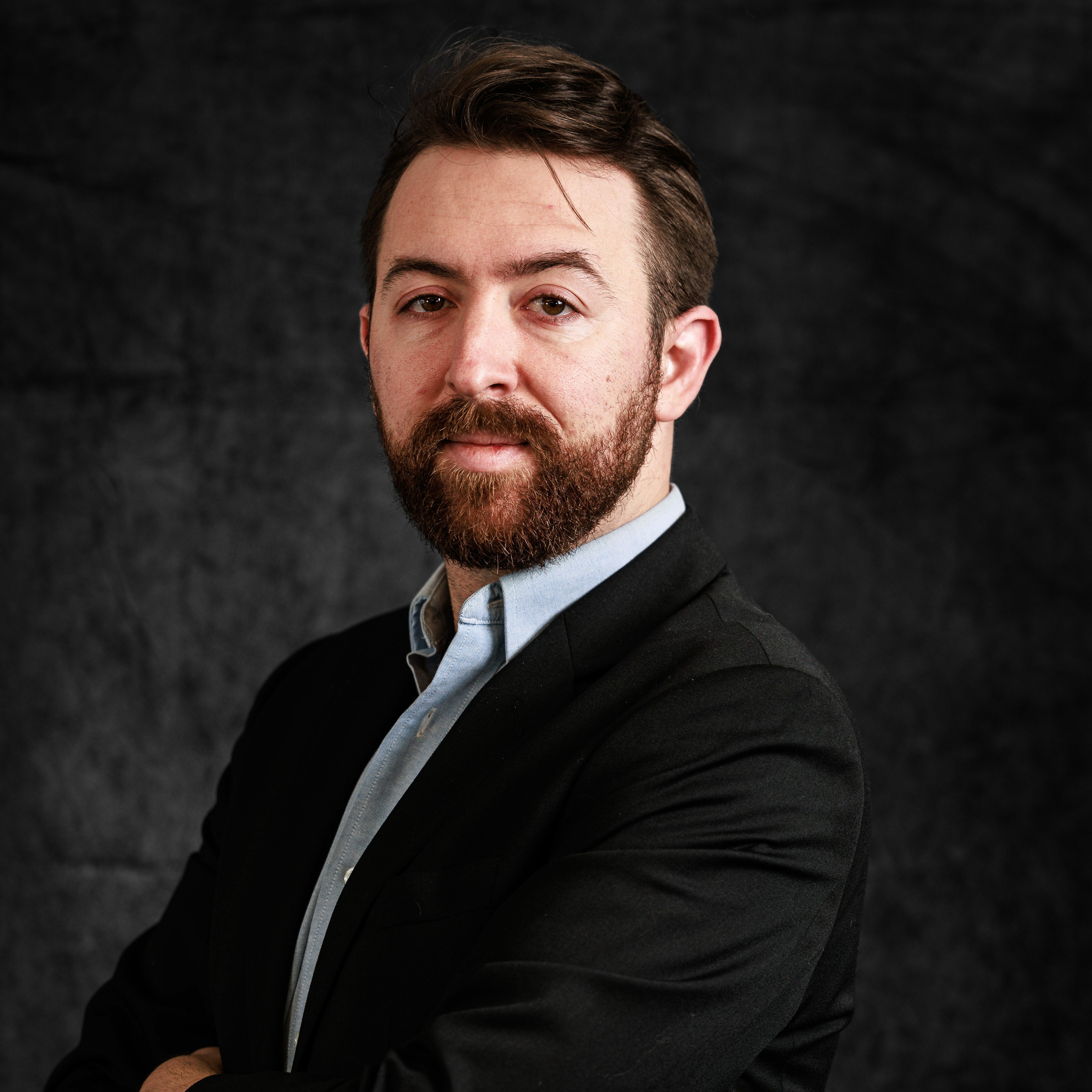 A handsome man with a beard in a suit, posing confidently against a dark background.