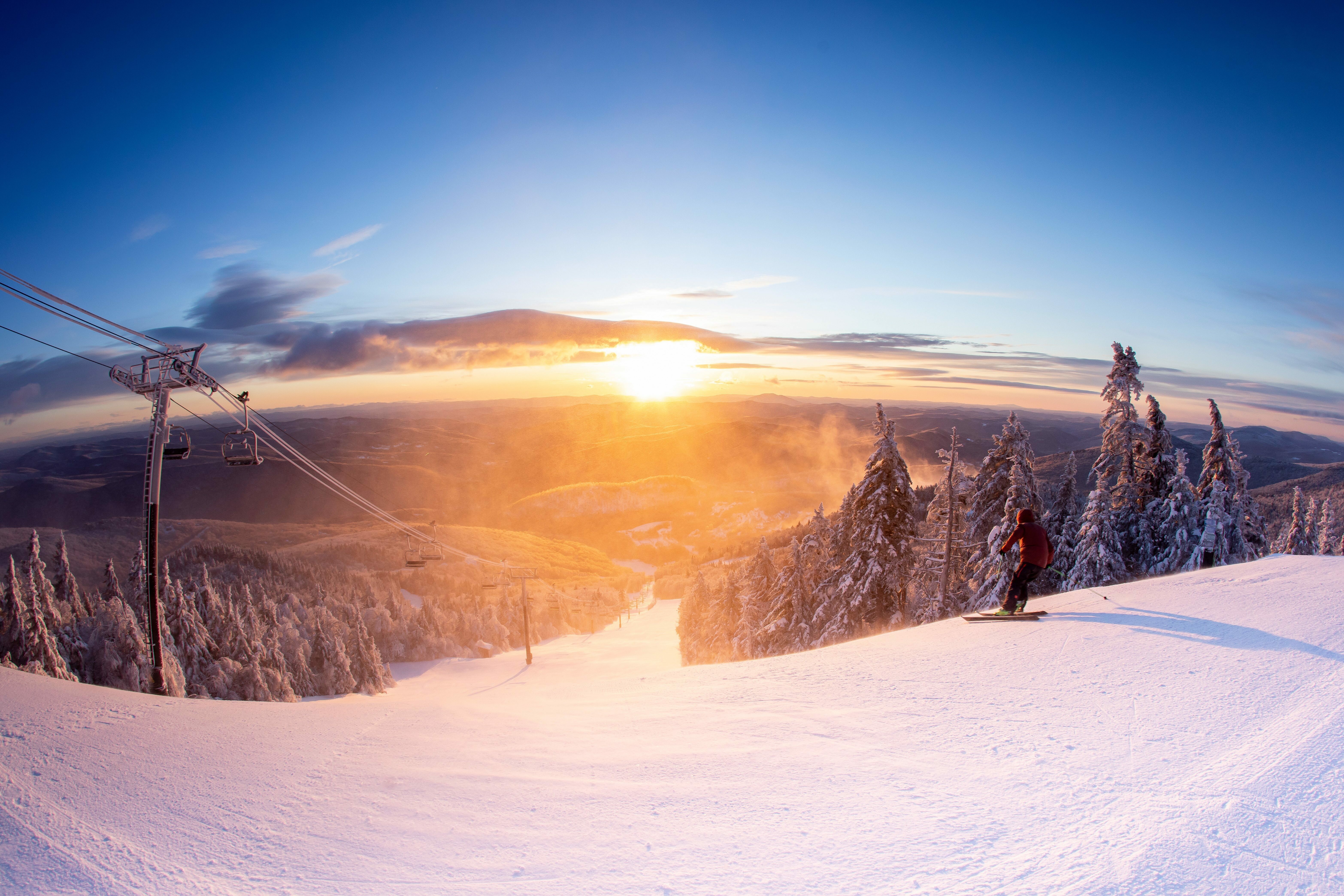 Skier on snowy slope at sunset, with lift and trees in the background.