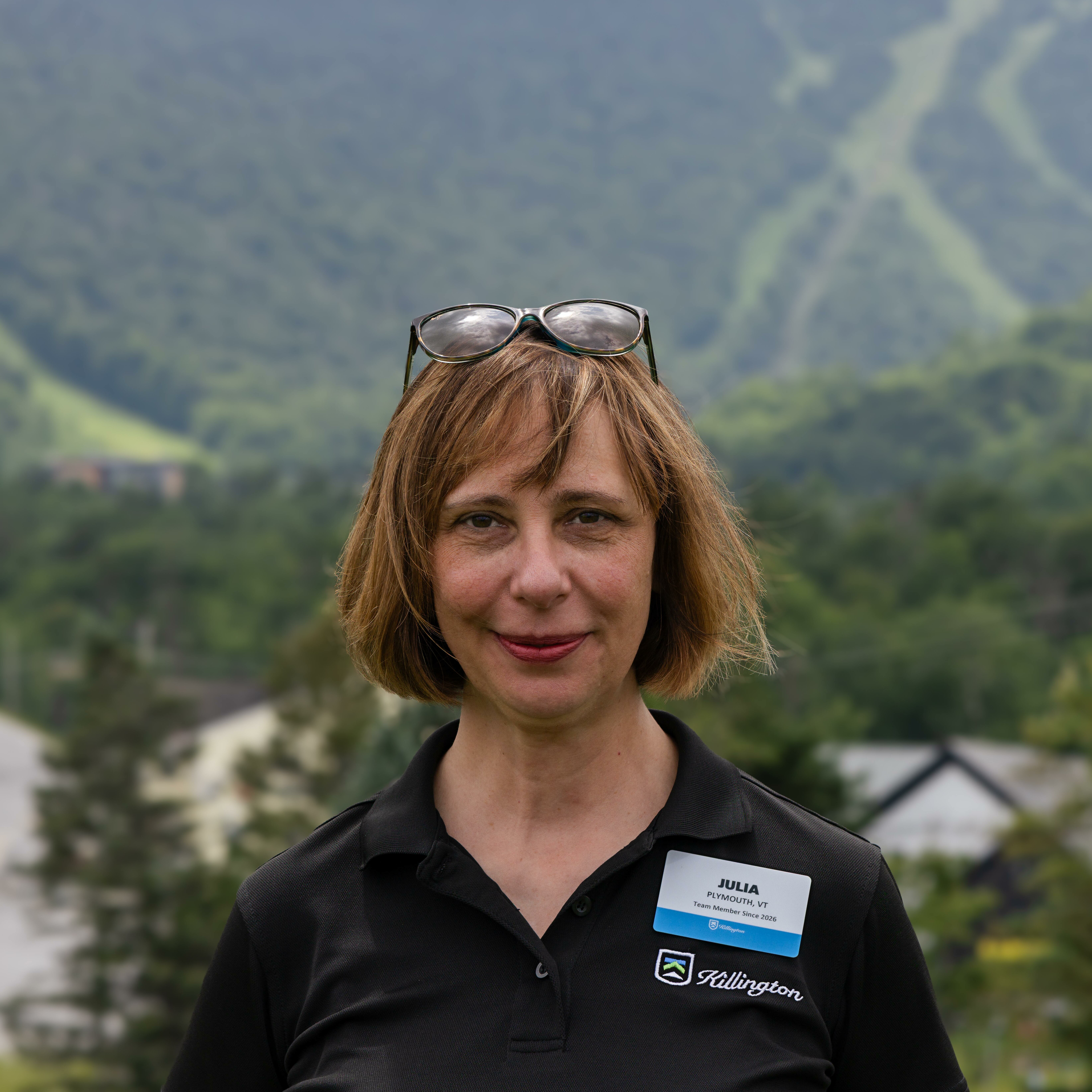 Woman with glasses on her head, wearing a name tag, with mountains in the background.
