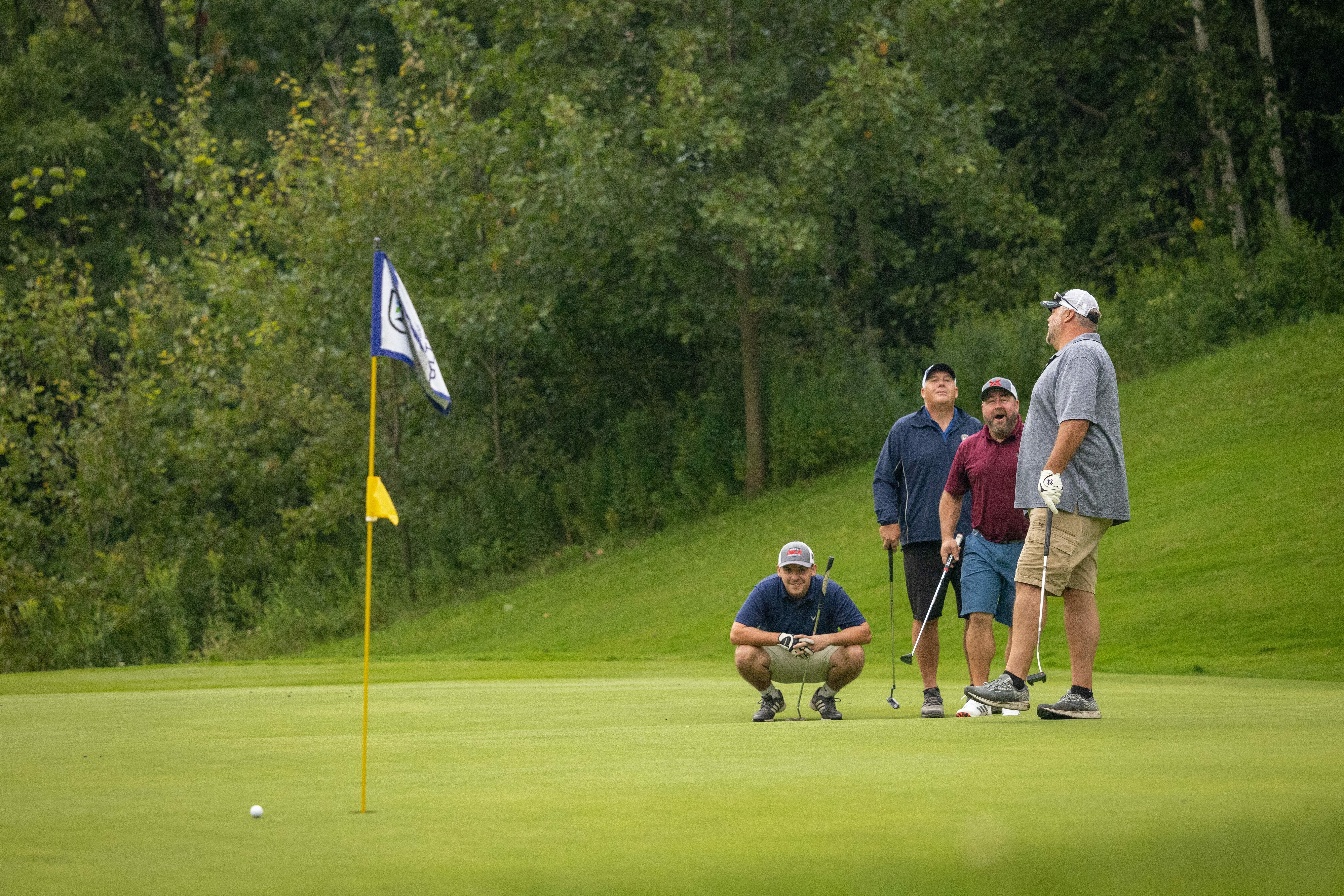 Four golfers observe a putt on the green, with a flag in the background.