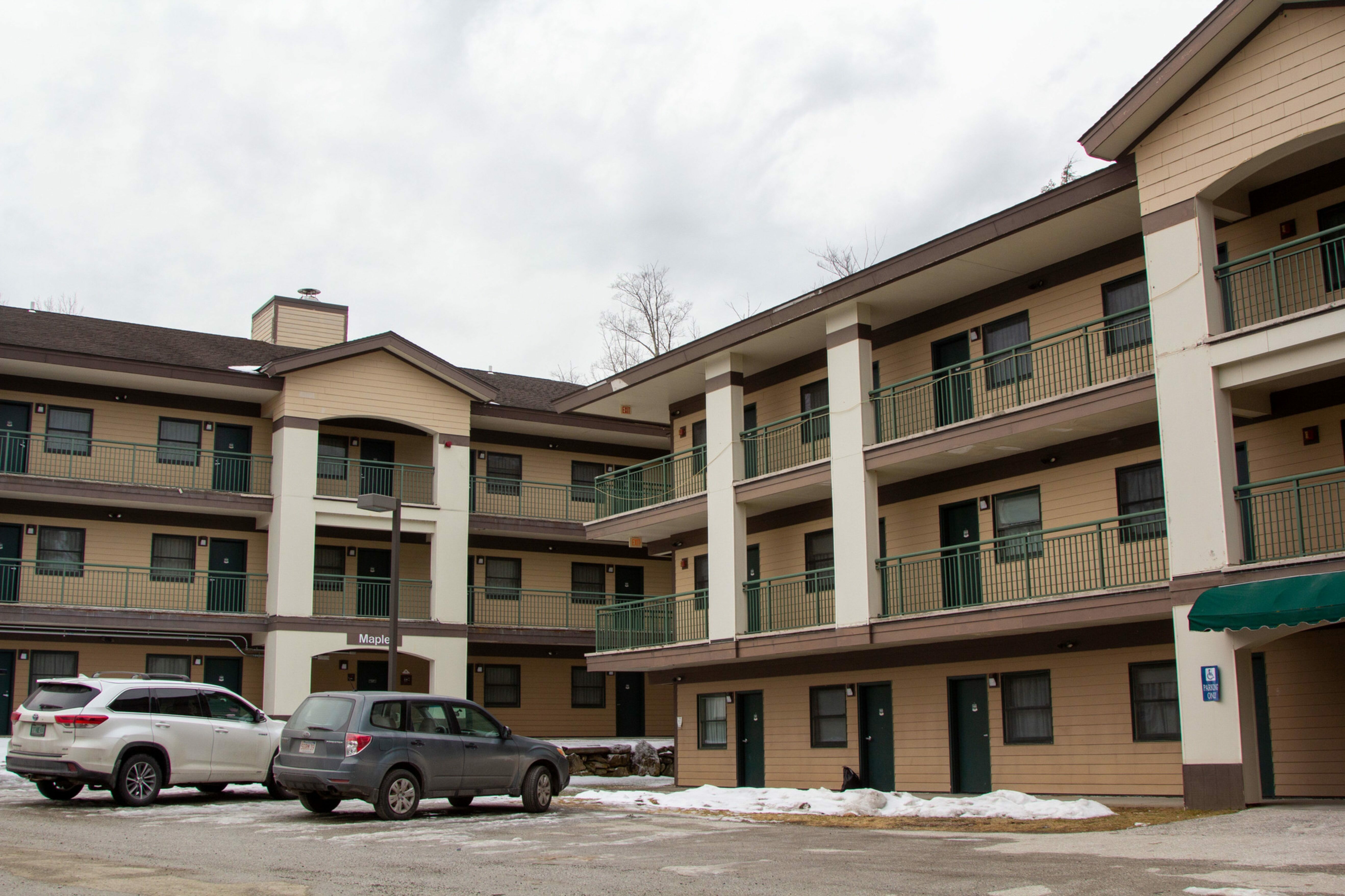 Three-story lodging building with balconies and parked vehicles outside.
