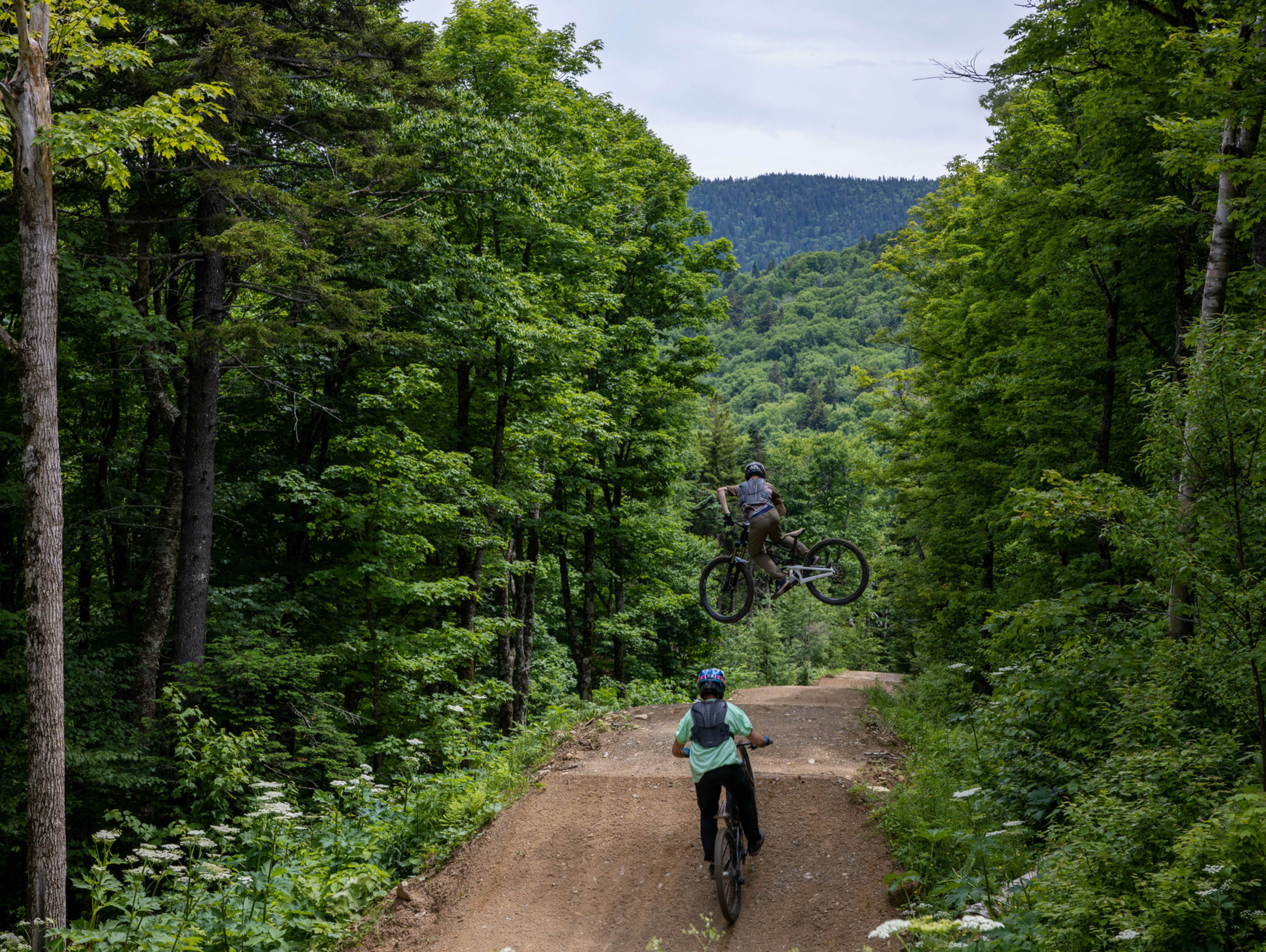 Two mountain bikers on a forested trail; one performs a jump over the other.