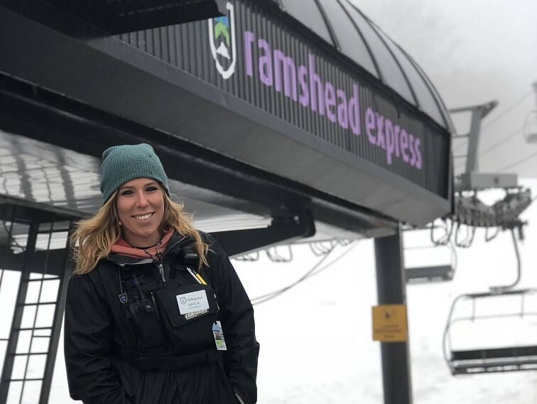 A smiling woman in winter attire stands under a ski lift sign that says "ramshead express."