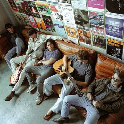 Five musicians relax on a couch, surrounded by colorful concert posters, with guitars in hand.