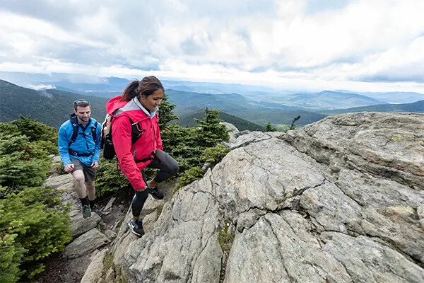 Two hikers navigate rocky terrain on a mountain with expansive green valleys in the background.