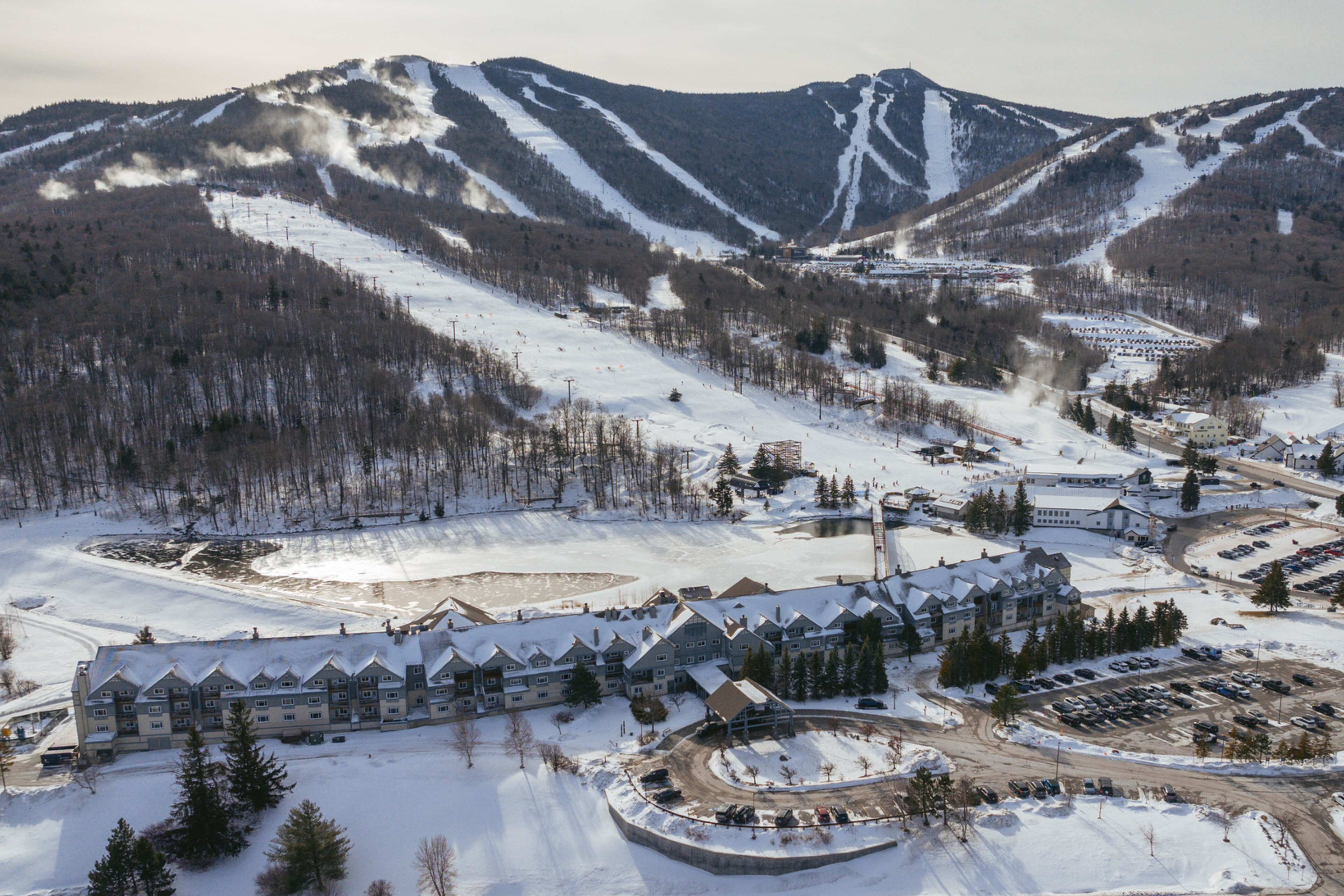 A snowy landscape featuring ski slopes, a resort, and a frozen pond in the foreground.