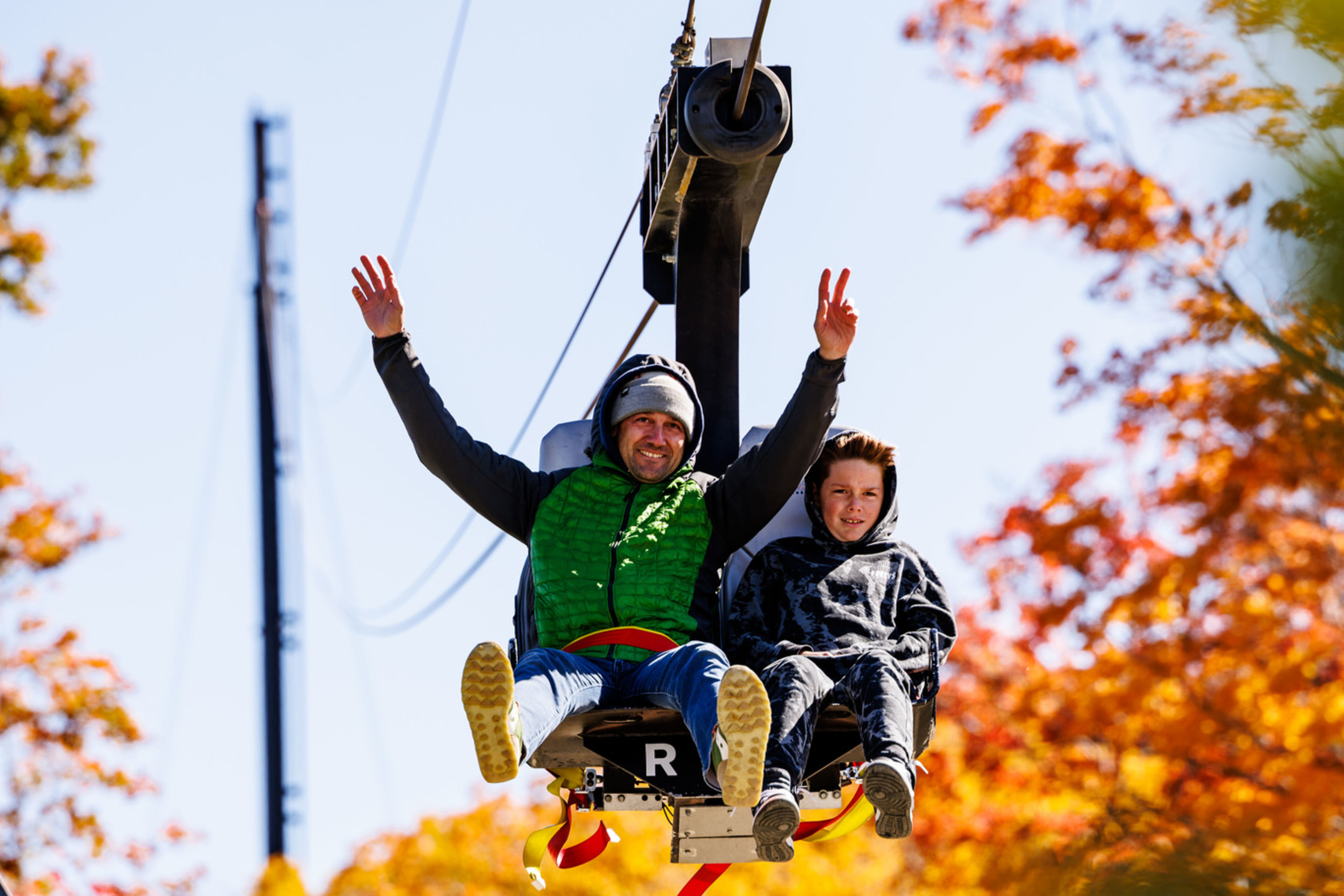 A man and a boy joyfully ride a ski lift with colorful autumn leaves in the background.